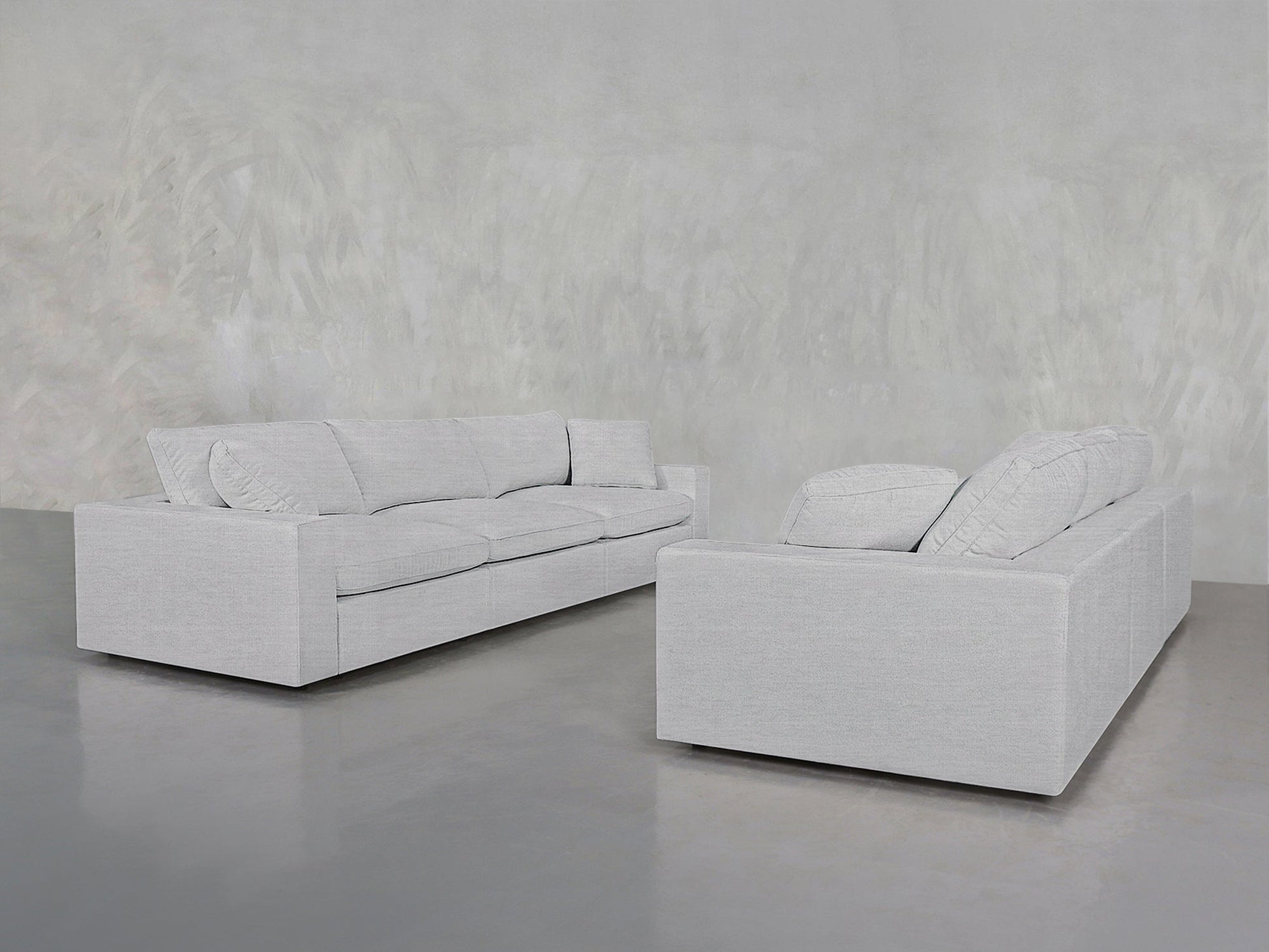 Two contemporary 3-3 Sofa Set pieces in light gray, featuring boxy shapes and plush cushions, are arranged opposite each other in a minimalist room with smooth gray concrete flooring and a textured gray wall.