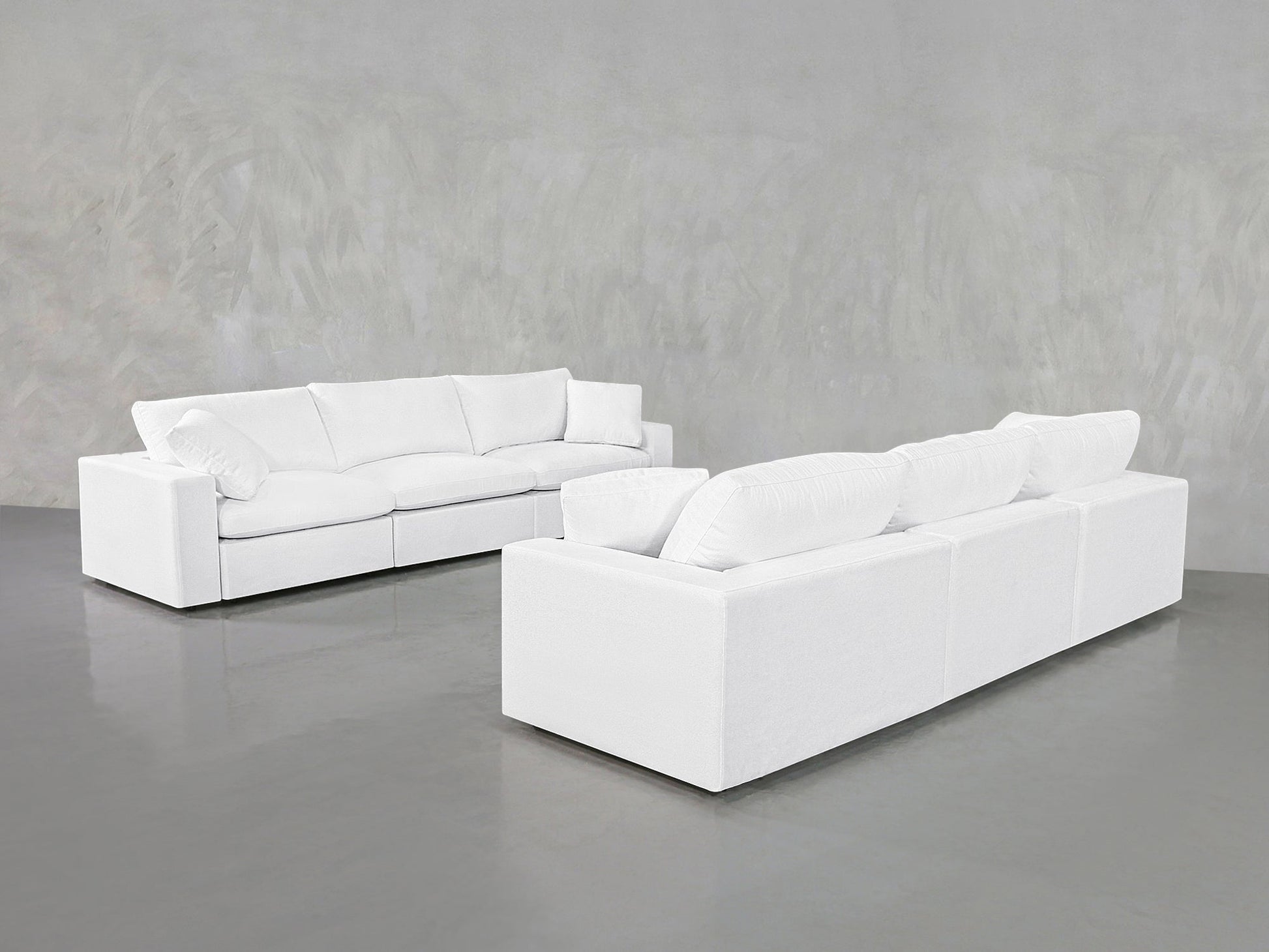Two stylish 3-3 Sofa Sets with plush cushions stand on a shiny gray floor against a light gray textured wall, creating a minimalist atmosphere.