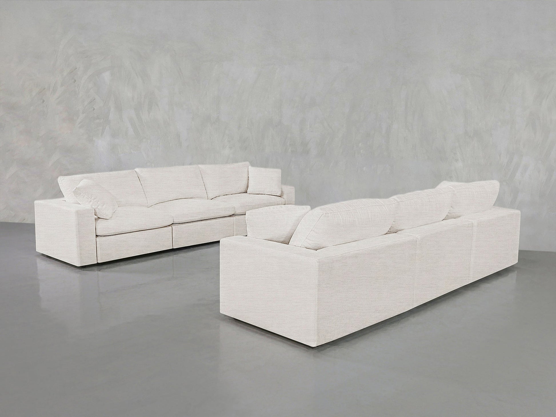 Two modern 3-3 Sofa Sets in light beige are arranged in an empty room with a smooth gray floor and textured light gray walls. These sofas feature plush cushions and a minimalist design.
