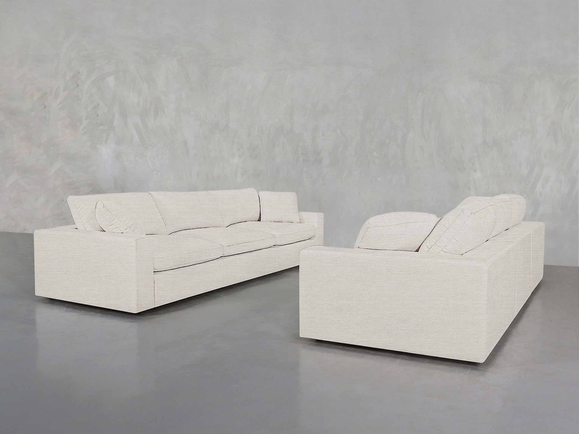 Two 3-3 Sofa Set pieces in light beige with clean lines and plush cushions are placed against a gray wall and polished concrete floor, creating a minimalist look.