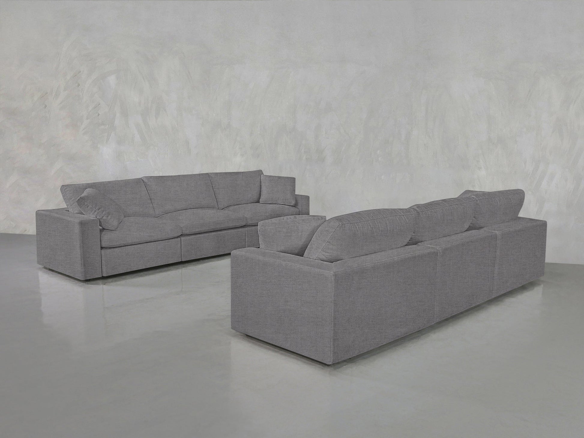 The 3-3 Sofa Set, featuring clean lines and plush cushions in modern gray fabric, is arranged in an empty room with a light gray textured wall and polished concrete floor.