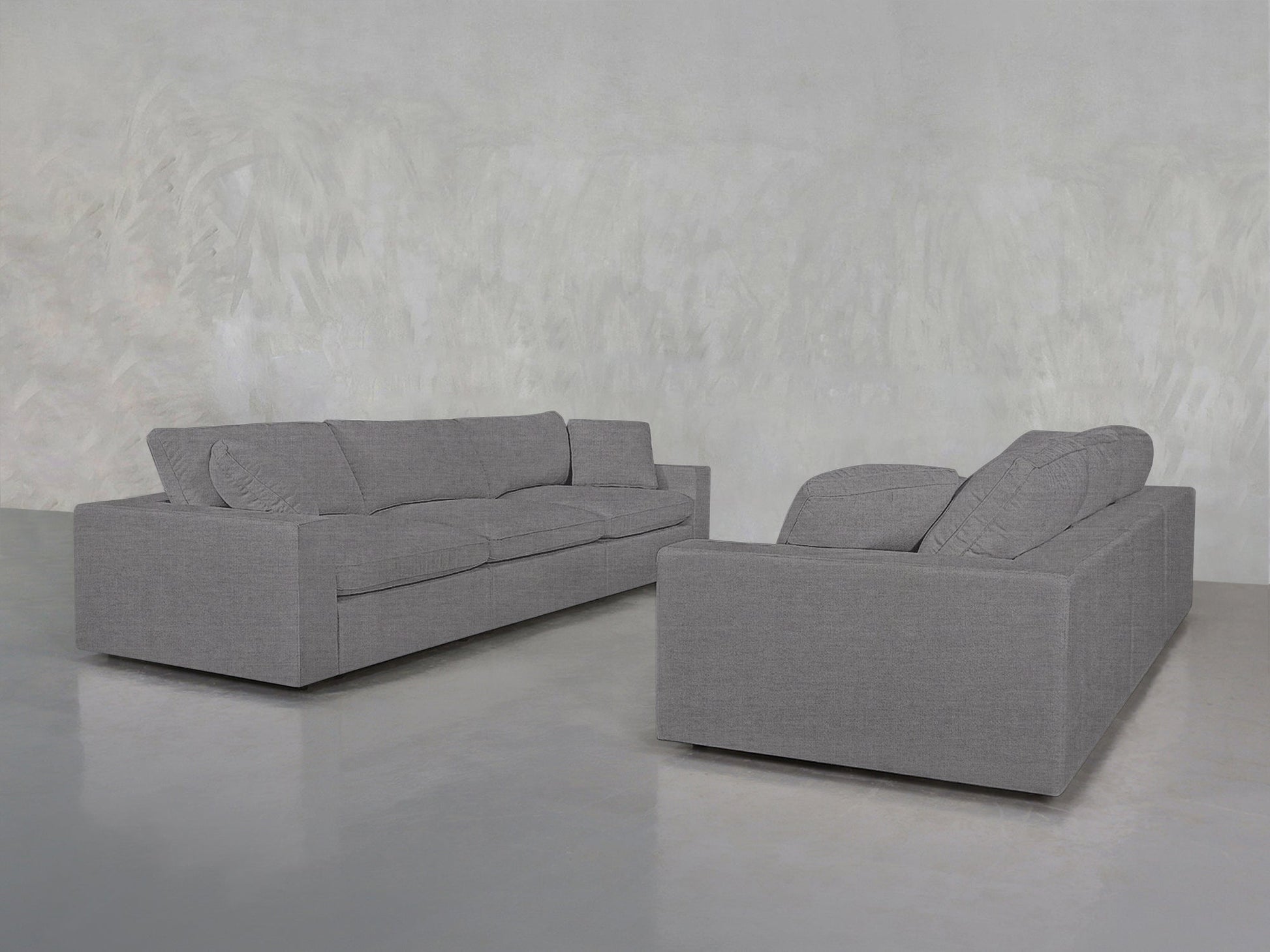 Two 3-3 Sofa Set pieces in modern grey, featuring clean lines and plush cushions, are arranged in an empty room with a light grey textured wall and a polished concrete floor.