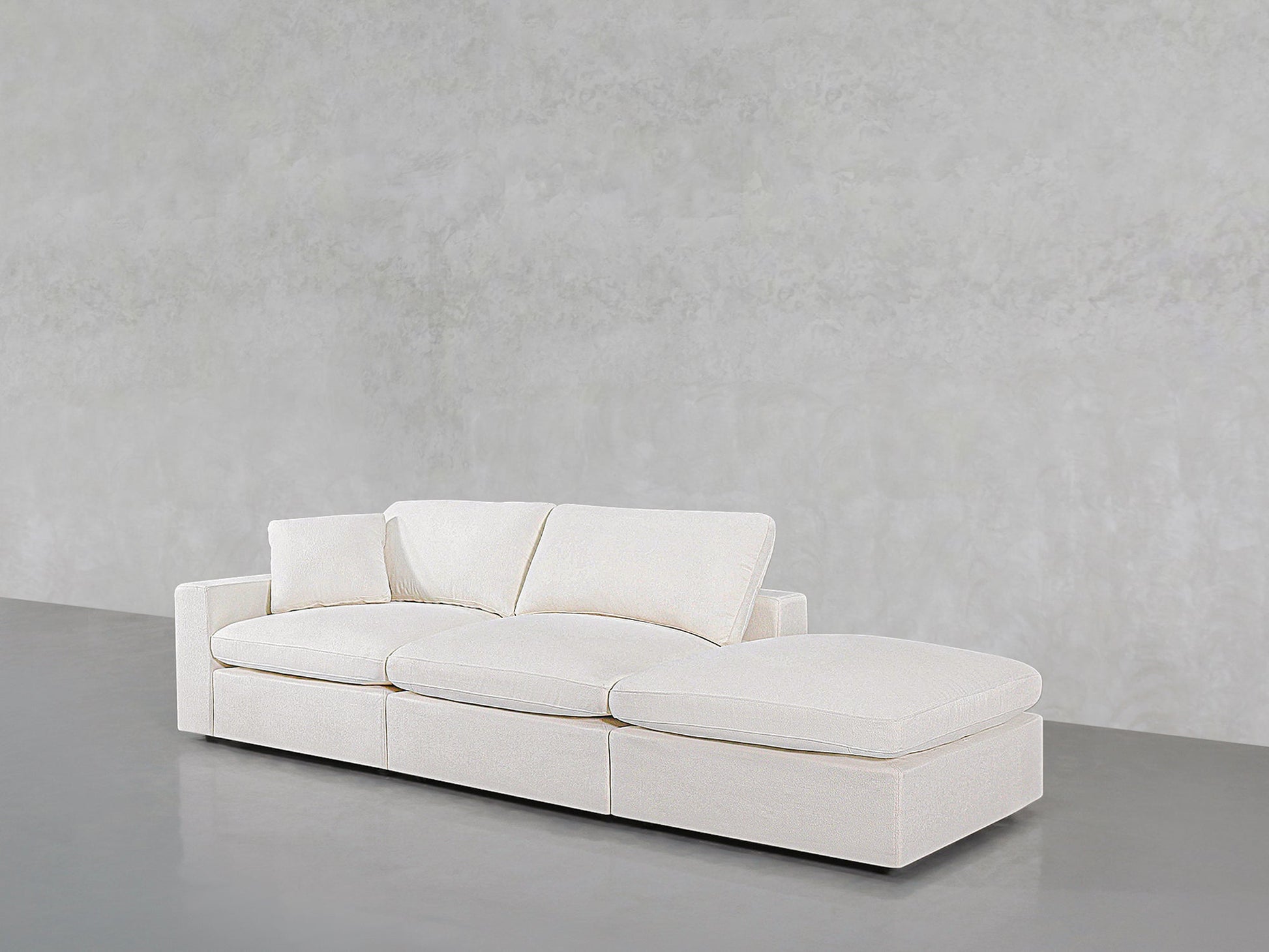 The 3-Seat Modular Lounger Sofa features white upholstery, three seat cushions, and a right-side chaise lounge, set against a light gray textured wall and smooth gray floor.