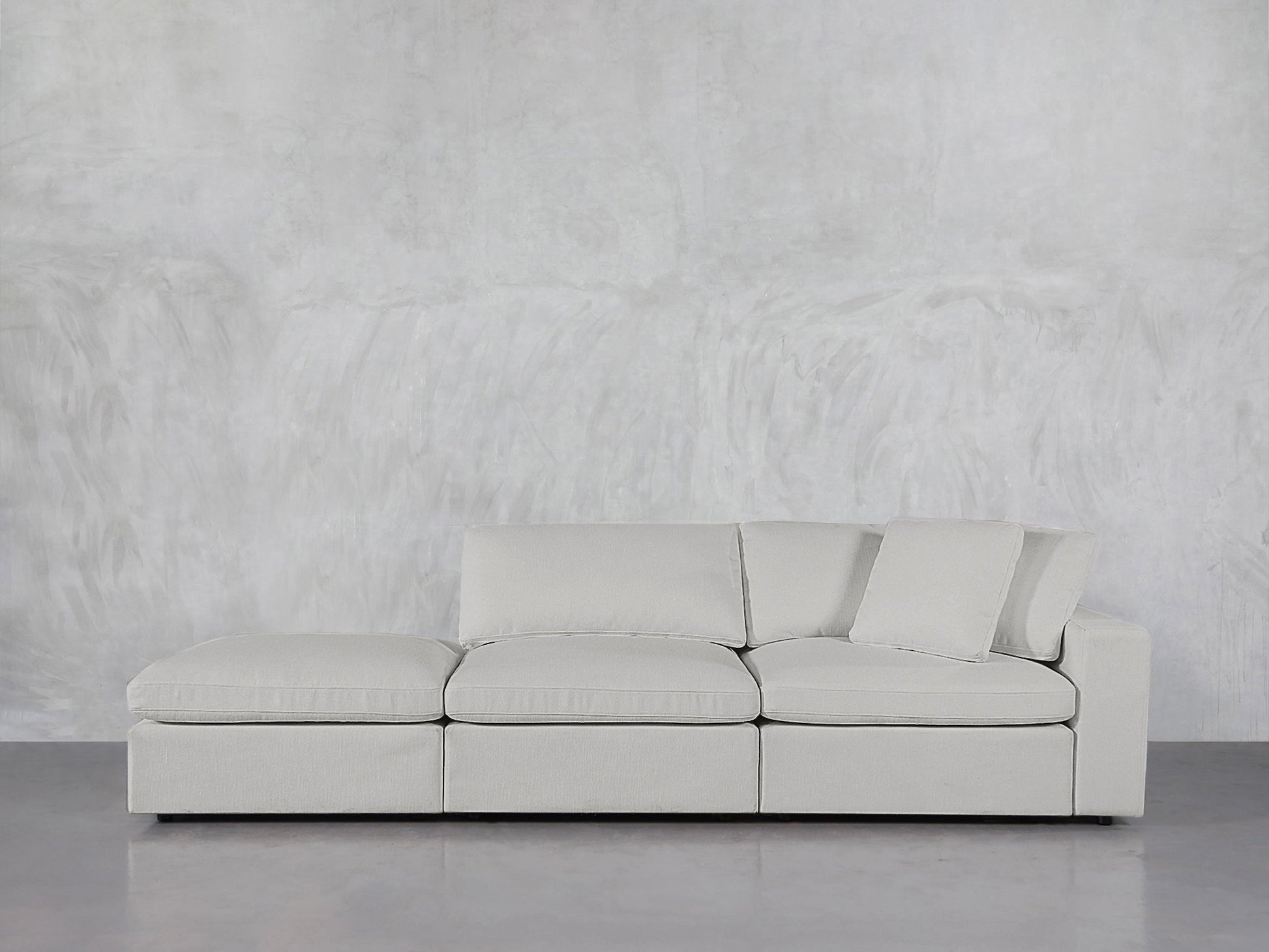 The 3-Seat Modular Lounger Sofa in light gray features clean lines and loose cushions, set against a textured gray wall on a polished floor. One back cushion is tilted against the armrest for a relaxed look.