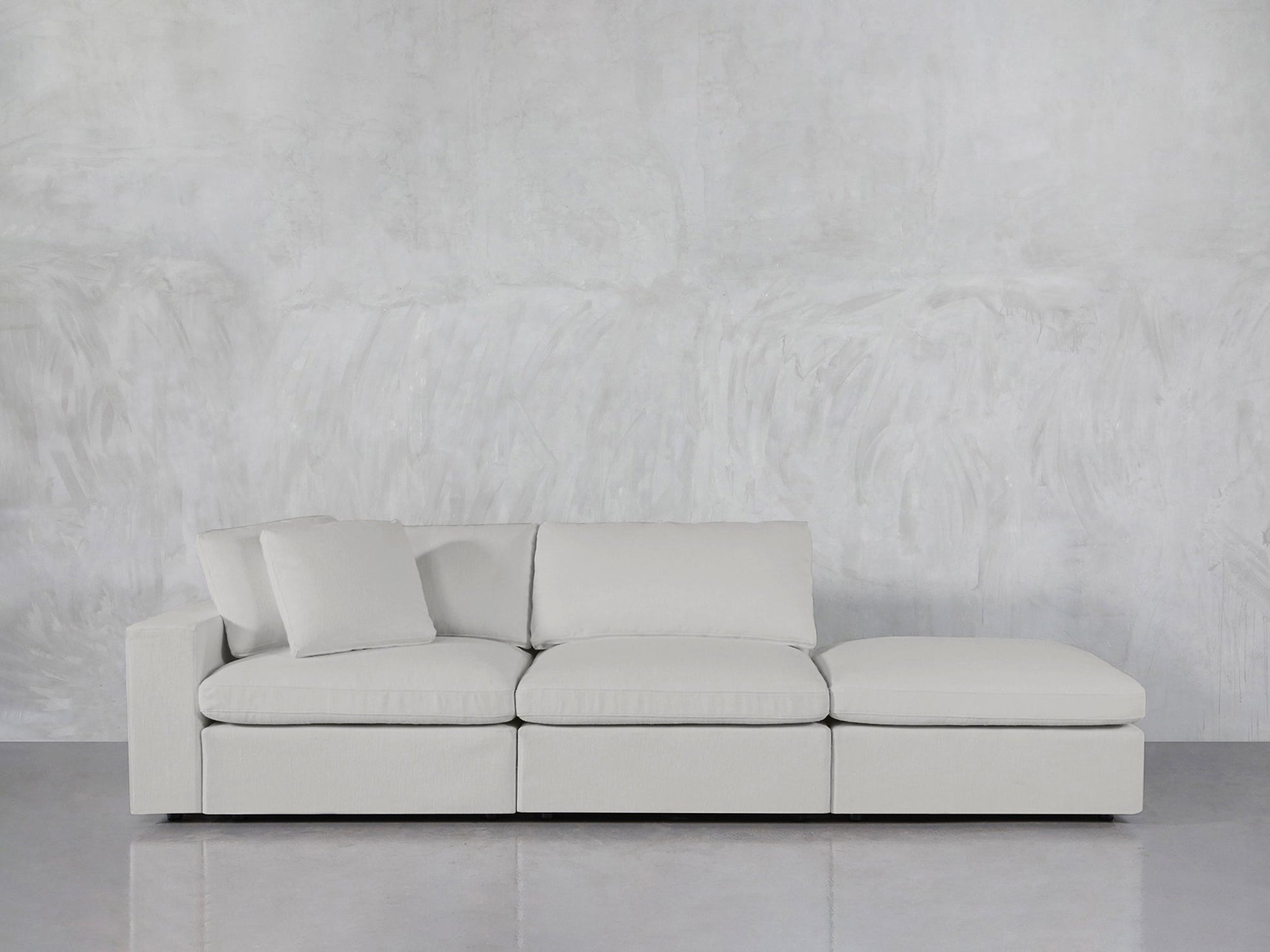 The 3-Seat Modular Lounger Sofa in light gray, with three cushions and a chaise lounge, is set against a textured gray wall on a polished gray floor. A single pillow rests on the left side of the sofa.