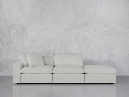 The 3-Seat Modular Lounger Sofa in light gray, with three cushions and a chaise lounge, is set against a textured gray wall on a polished gray floor. A single pillow rests on the left side of the sofa.