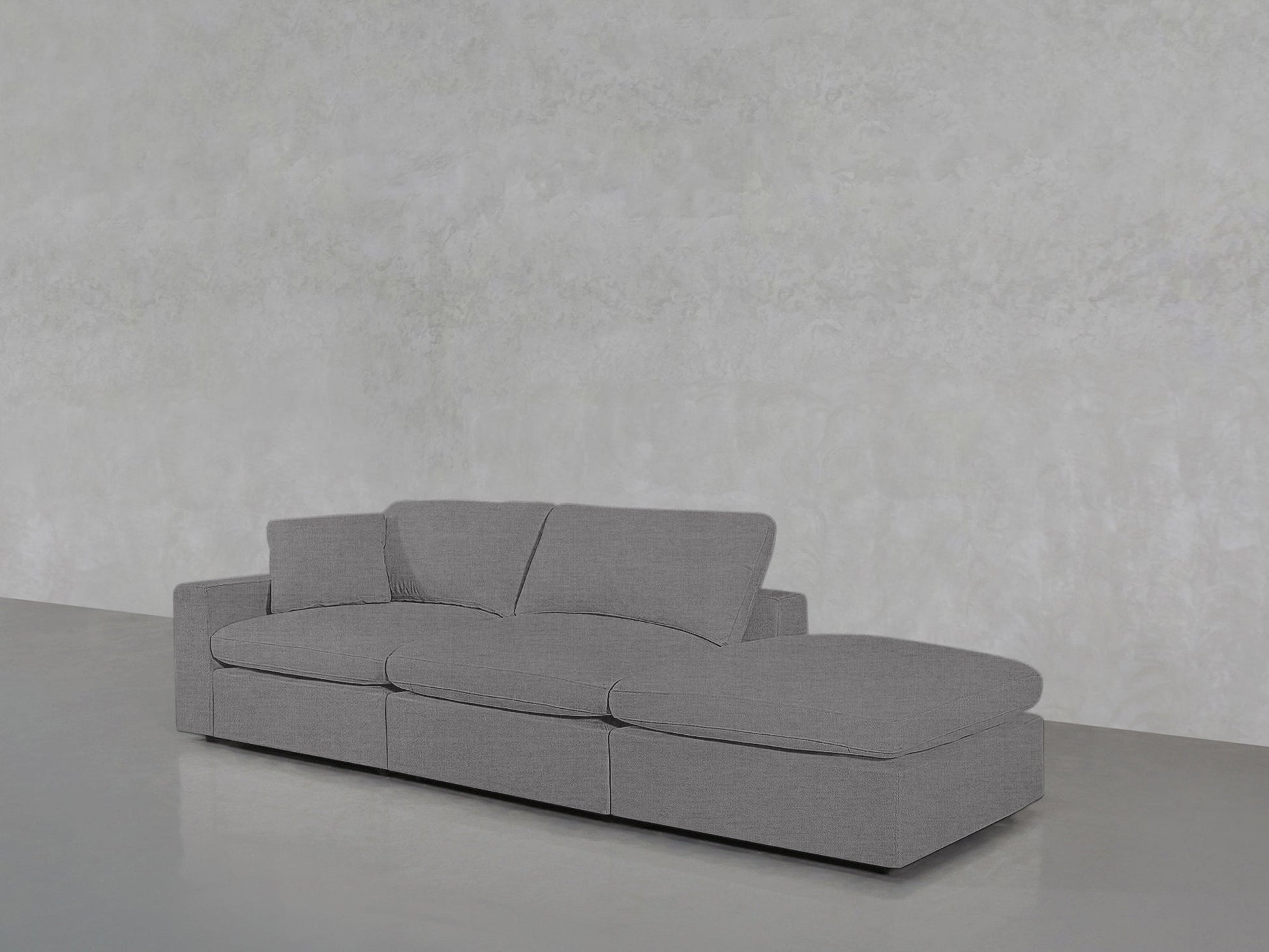 A 3-Seat Modular Lounger Sofa with plush gray cushions is placed on a glossy light gray floor against a textured light gray wall in a minimalist, empty room.