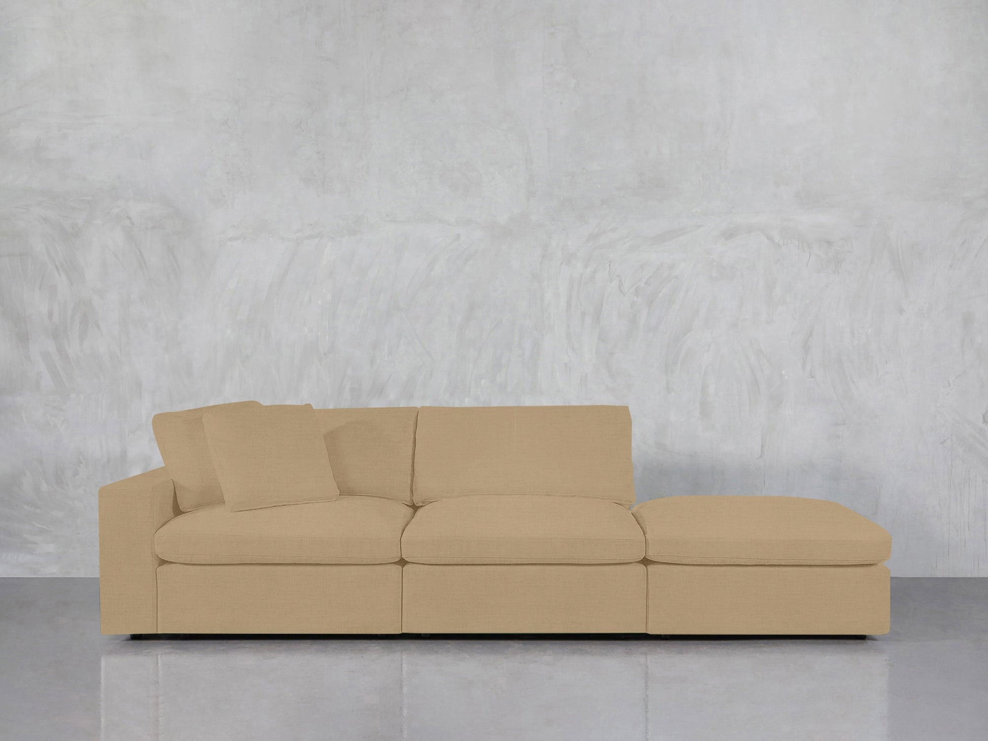 A 3-Seat Modular Lounger Sofa in beige, featuring an attached chaise lounge and two matching cushions on the left, is set against a light gray textured wall with a reflective floor beneath it.