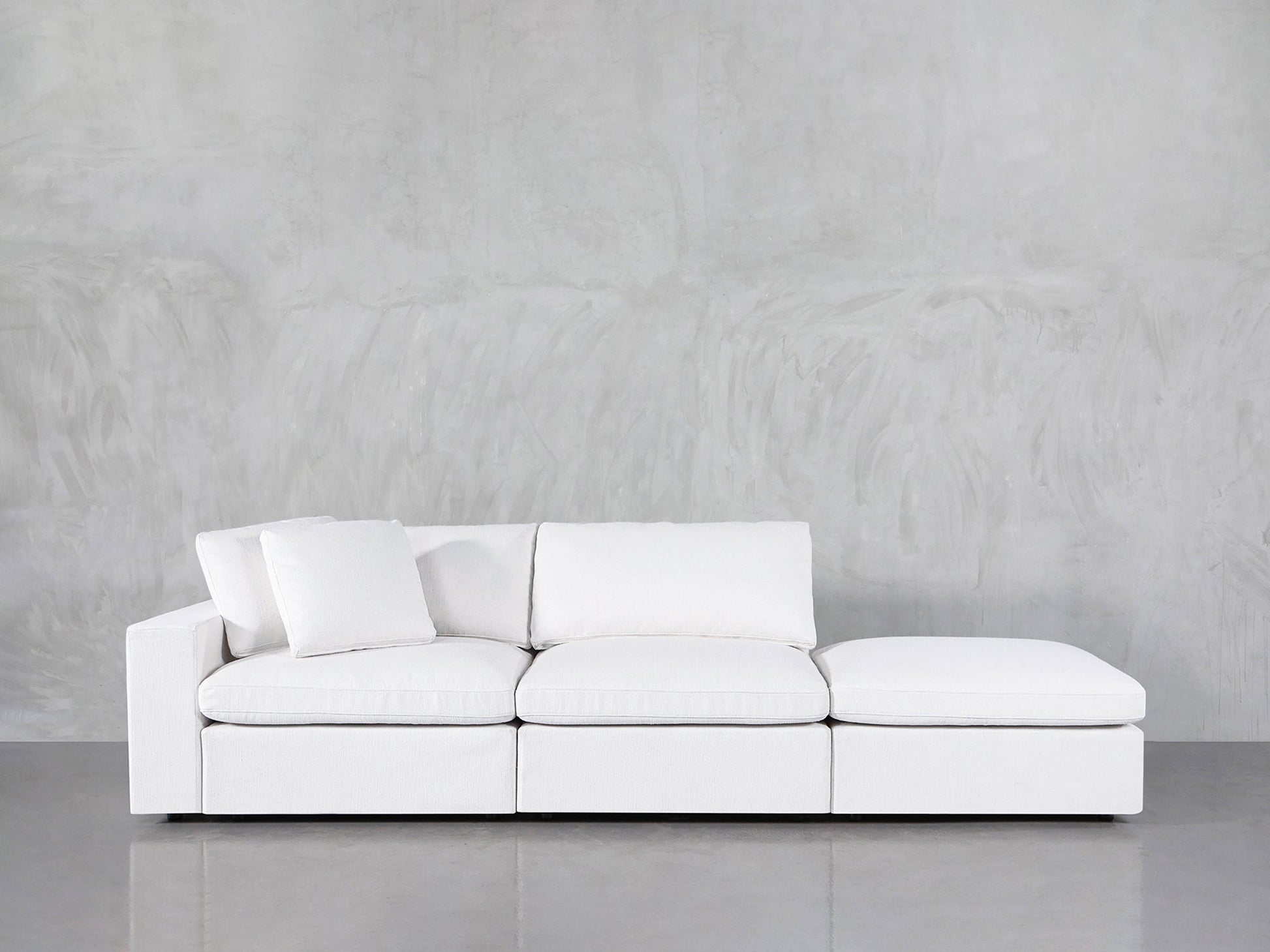 A 3-Seat Modular Lounger Sofa in white with three seat cushions and an ottoman is placed before a light gray textured wall and a reflective gray floor. One white pillow is positioned on the left side of the sofa.