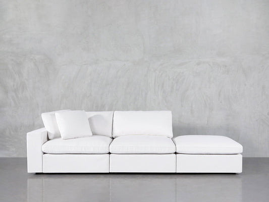 A 3-Seat Modular Lounger Sofa in white with three seat cushions and an ottoman is placed before a light gray textured wall and a reflective gray floor. One white pillow is positioned on the left side of the sofa.