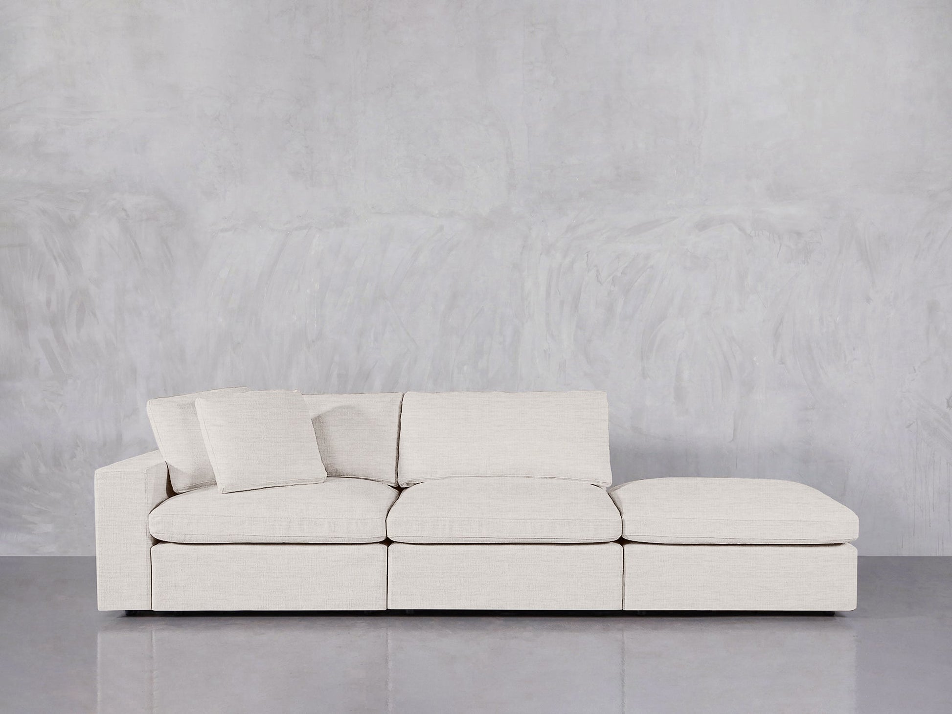 The 3-Seat Modular Lounger Sofa in light beige features three seats, a chaise lounge, and two matching cushions, displayed against a gray textured wall and reflective gray floor.