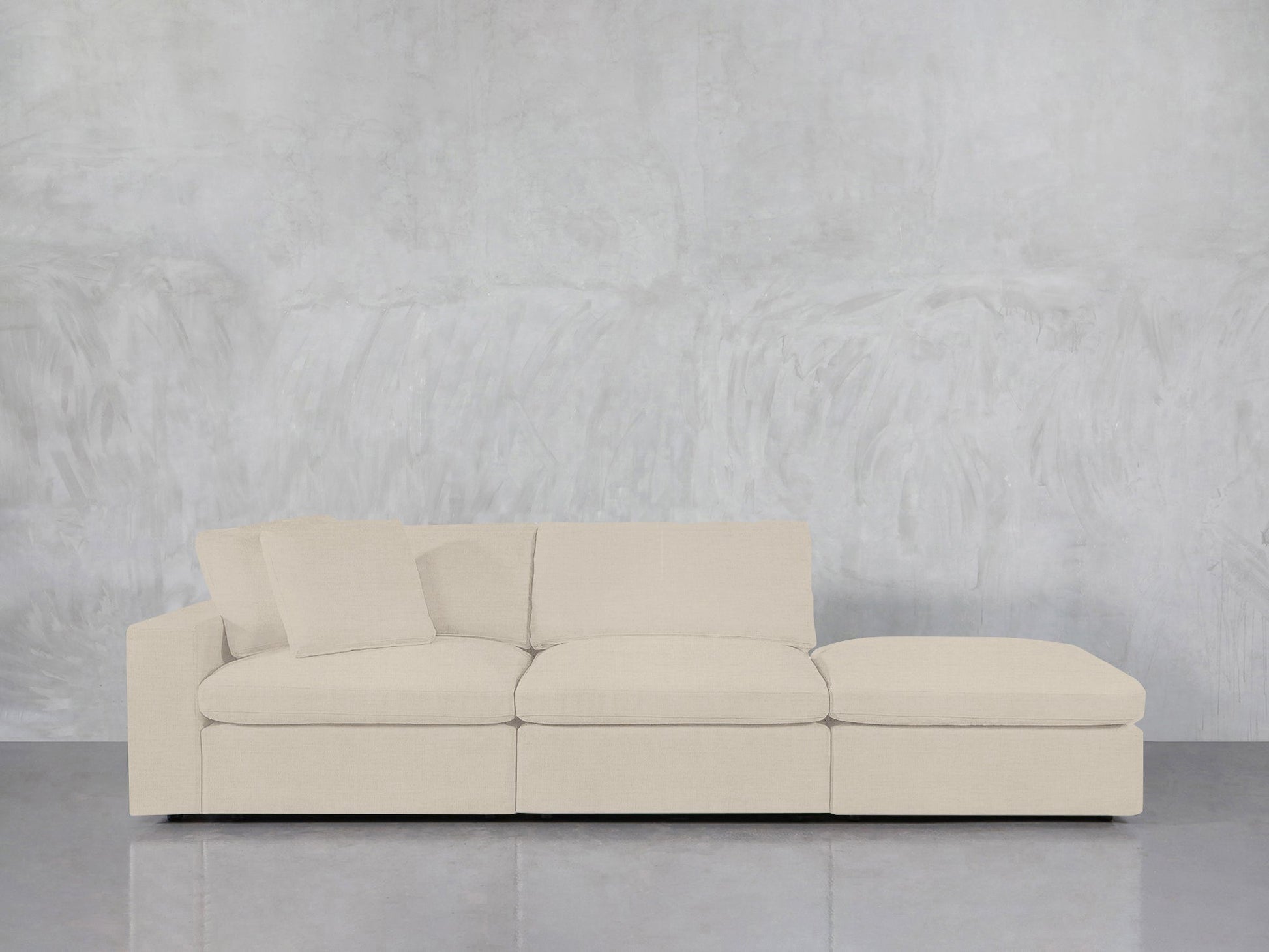 A 3-Seat Modular Lounger Sofa in light beige with two matching throw pillows is set against a textured light gray wall on a shiny gray floor.