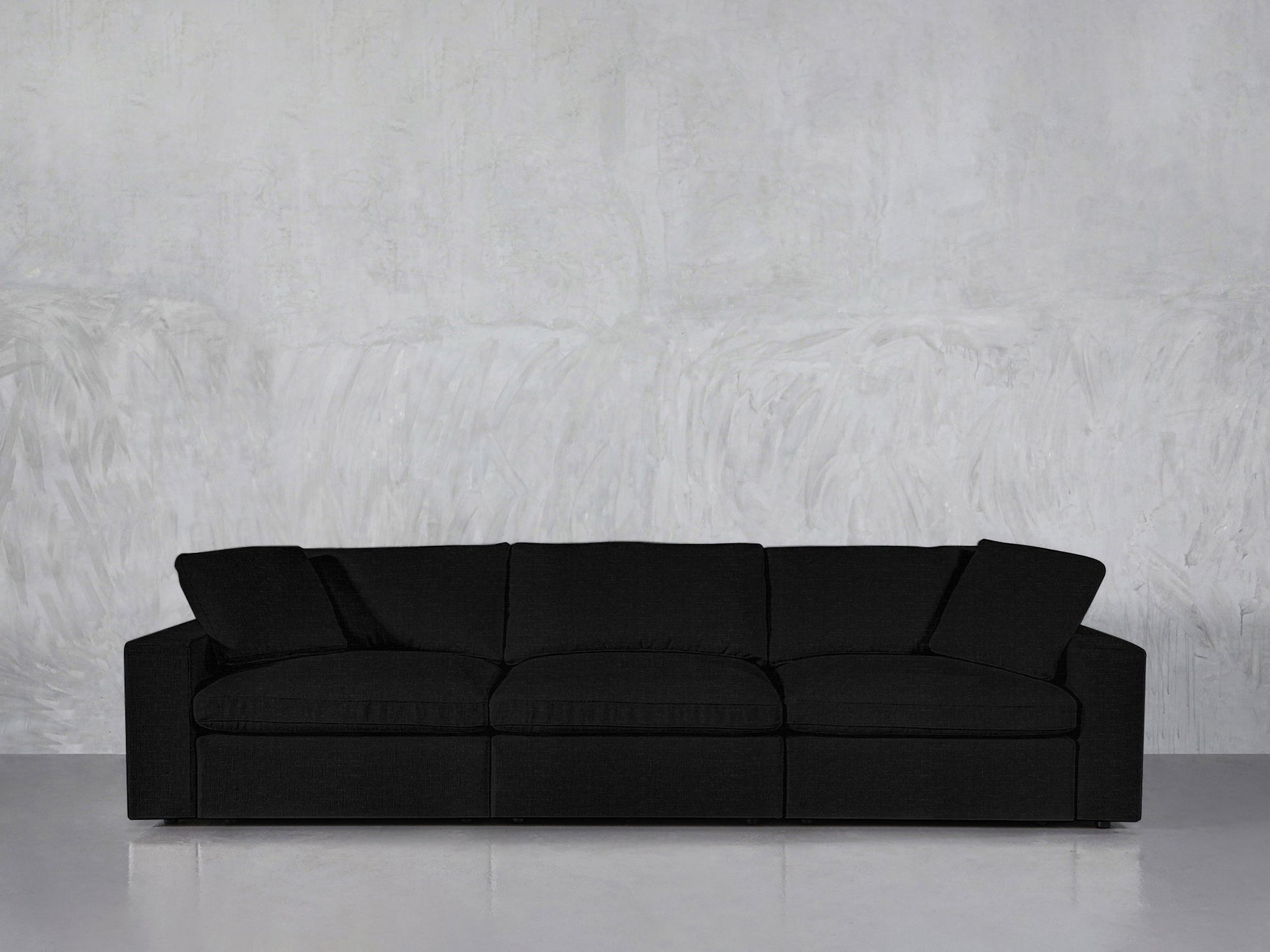 The 3-Seat Modular Sofa in modern black, featuring three seat cushions and two armrest pillows, is positioned against a light gray textured wall on a smooth, reflective floor.