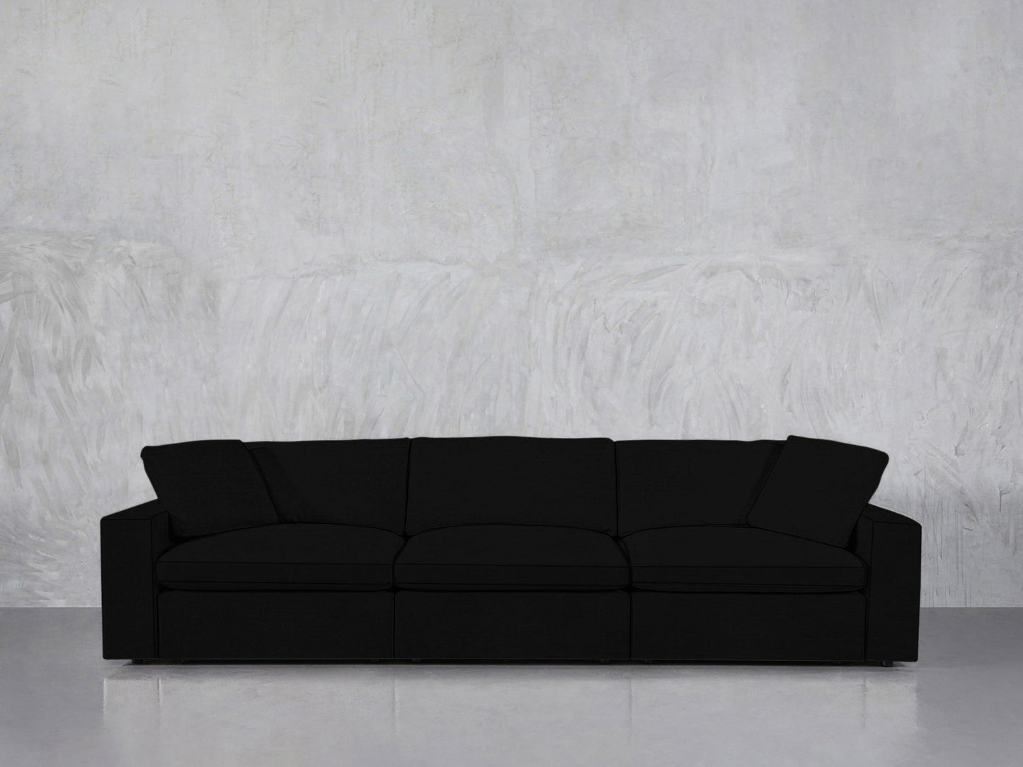 The 3-Seat Modular Sofa, in modern black with three cushions and two armrests, is set against a light gray textured wall on a smooth, reflective floor.