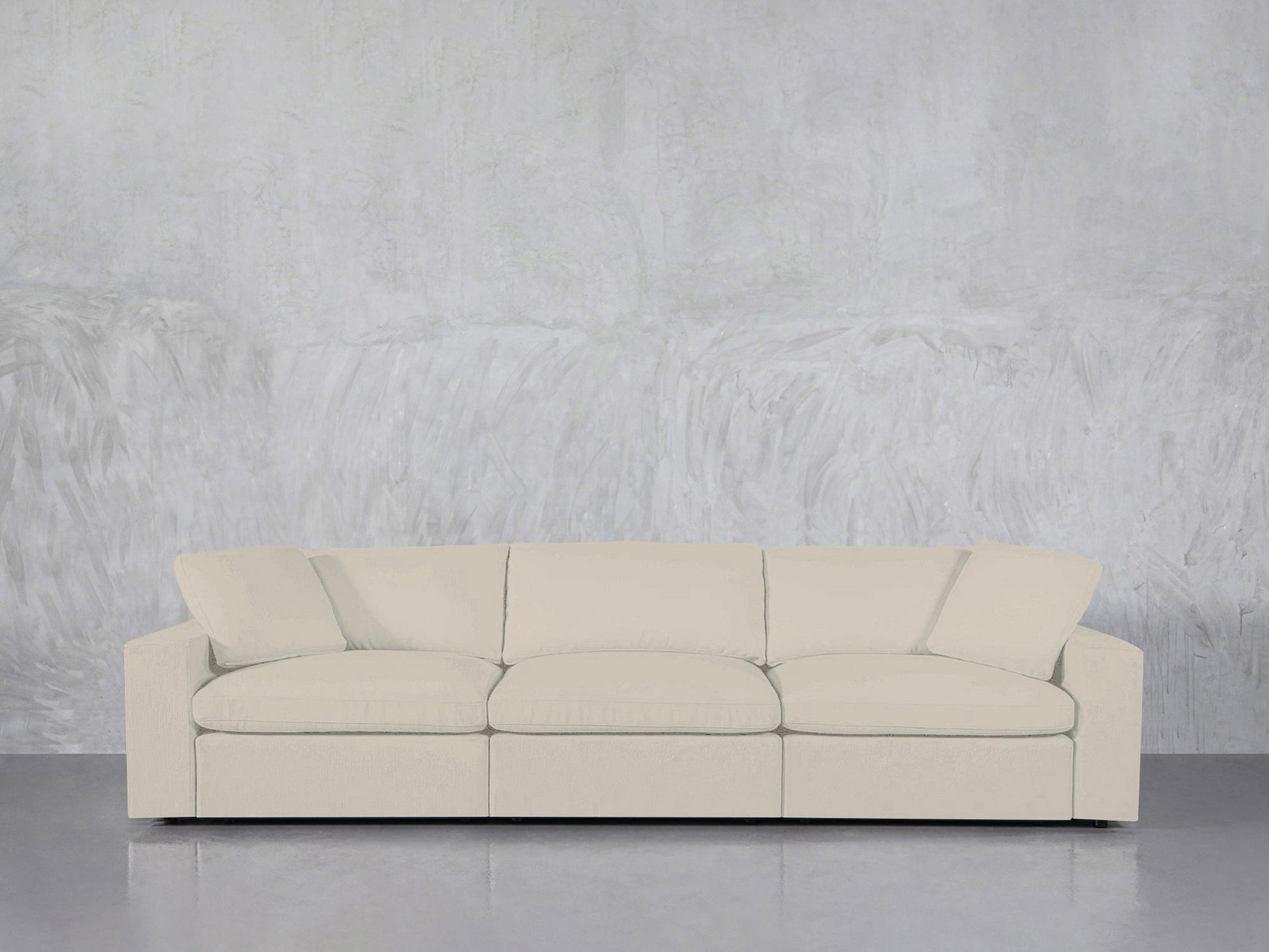 The 3-Seat Modular Sofa, in cream color with three seat cushions and an angled back cushion, is set against a textured light gray concrete wall and a smooth gray floor.