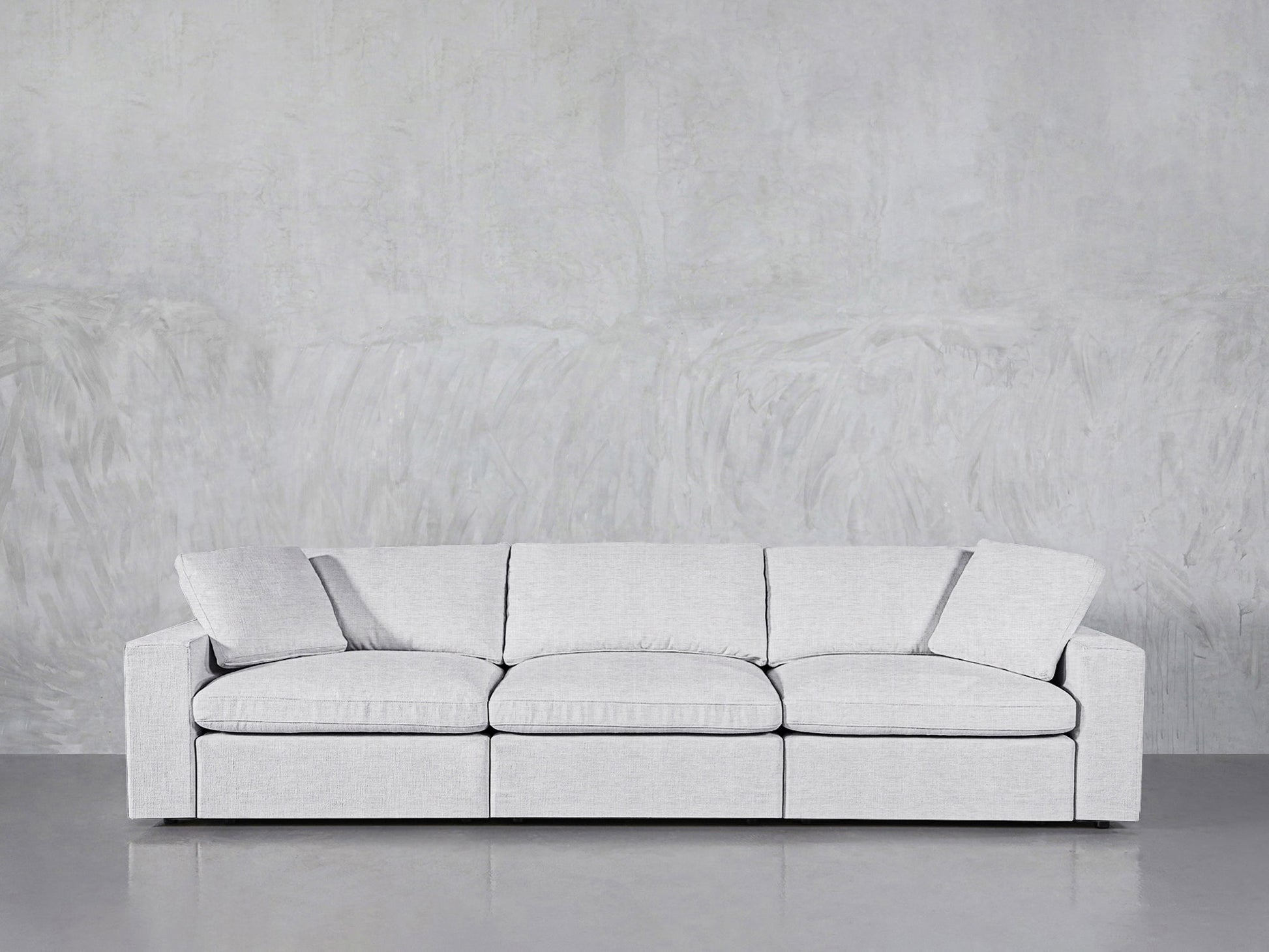 The 3-Seat Modular Sofa in light gray, featuring three seat cushions and a matching pillow, is placed against a textured gray wall and a smooth gray floor.