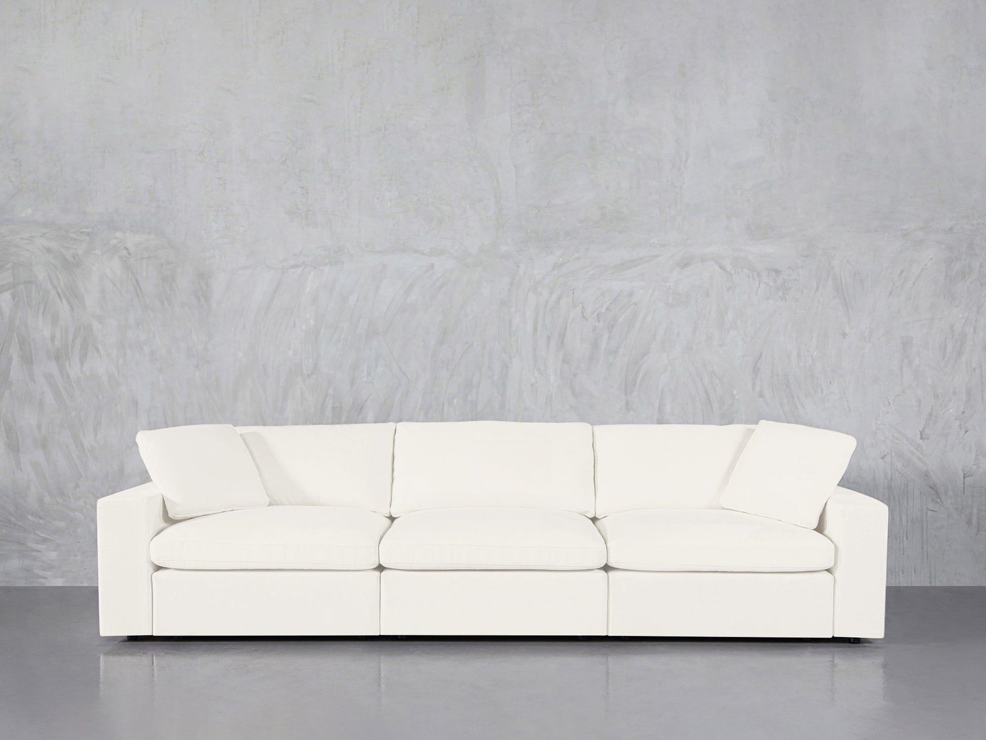 The 3-Seat Modular Sofa, featuring a modern white design with three seat cushions and two angled back pillows, is set against a light gray textured wall on a polished gray floor.