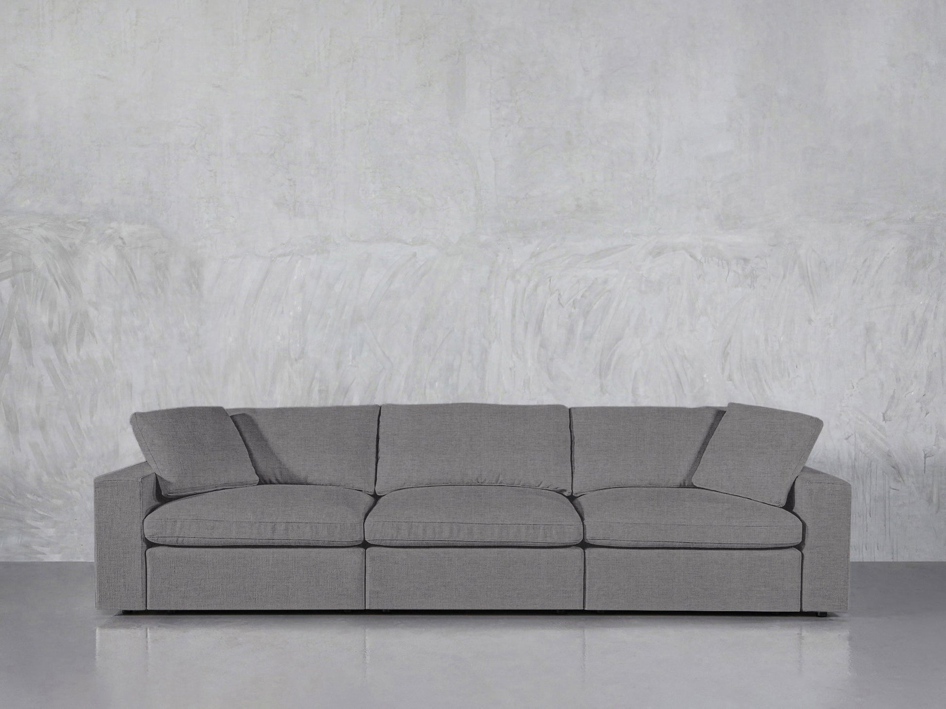 The 3-Seat Modular Sofa, upholstered in modern gray fabric with three seat and back cushions plus slightly tilted armrests, rests against a textured light gray wall on a smooth, light gray floor.