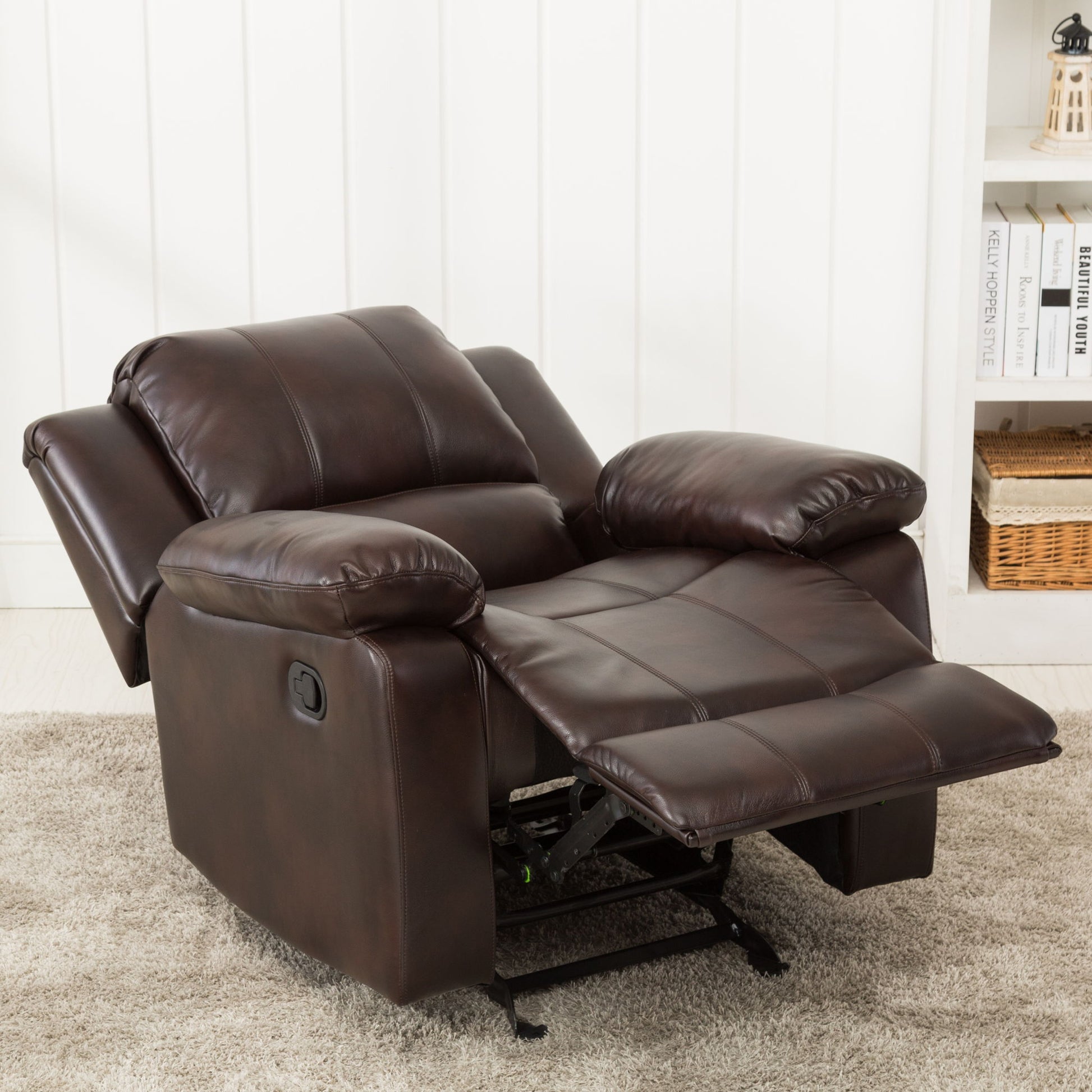 The Charlotte - Glider Recliner With Classic Style, in dark brown leather with an extended footrest, sits on a light carpet in a room featuring white paneled walls and shelves filled with books and baskets.