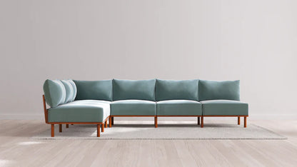 The Indoor Jack 6-Seater Open-Ends Corner Sectional in Balsa, featuring teal cushions and wooden legs, is set on a light-colored rug against a plain gray wall. The space maintains a minimalist aesthetic with its light hardwood flooring.