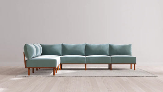 The Indoor Jack 6-Seater Open-Ends Corner Sectional in Balsa, featuring teal cushions and wooden legs, is set on a light-colored rug against a plain gray wall. The space maintains a minimalist aesthetic with its light hardwood flooring.