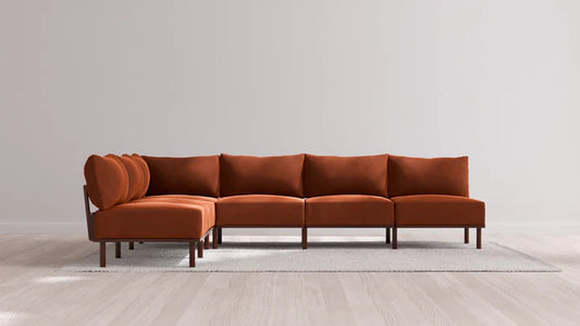 An Indoor Jack 6-Seater Open-Ends Corner Sectional in Balsa, featuring a modular orange-brown design, is arranged in an L-shape on a light gray rug. The setup is positioned against a plain, light-colored wall with a wooden floor.