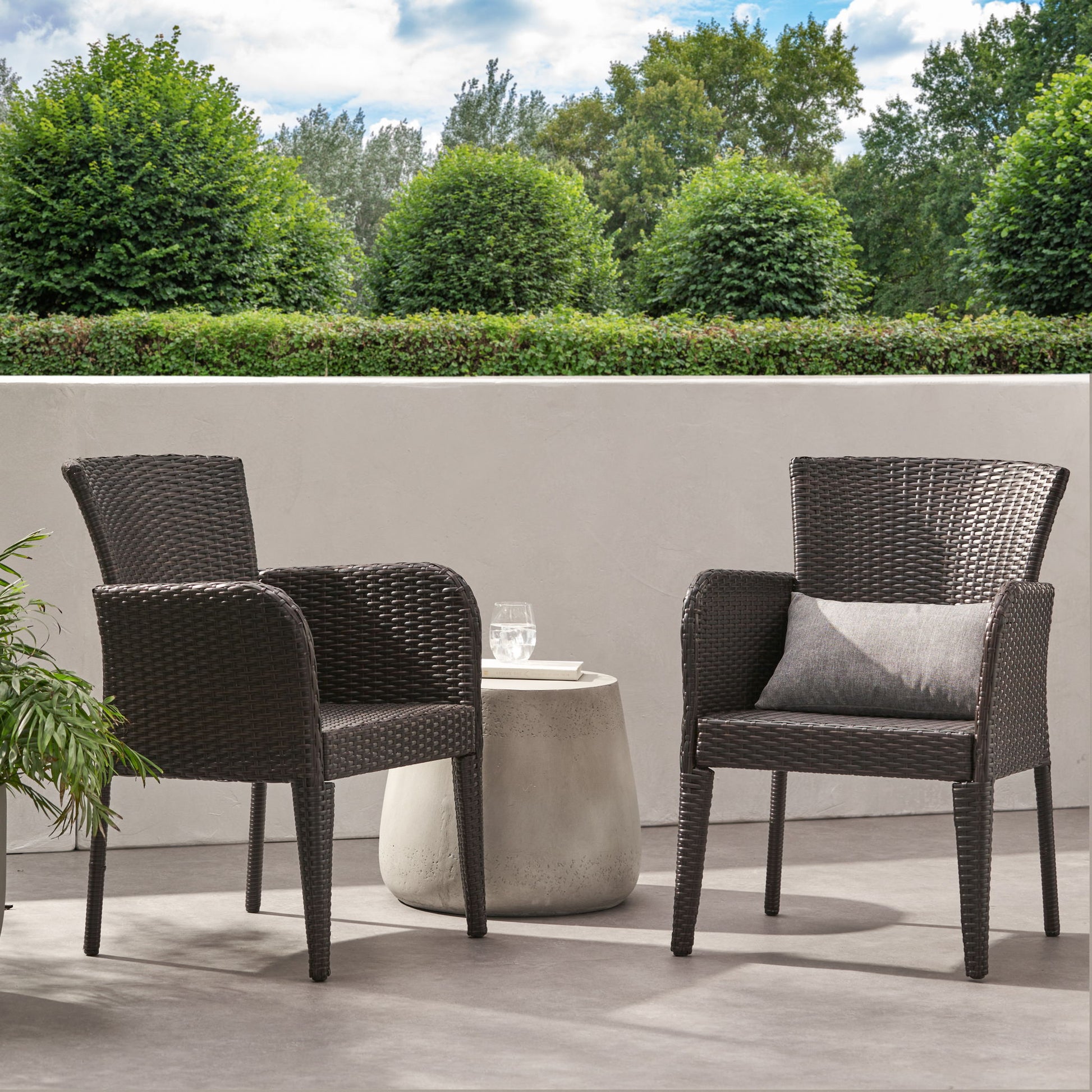 Two Anaya Outdoor Wicker Dining Armchairs with armrests and gray cushions are on a patio by a round table with a glass of water. Green bushes, trees, and a white wall stand in the background under partly cloudy skies.