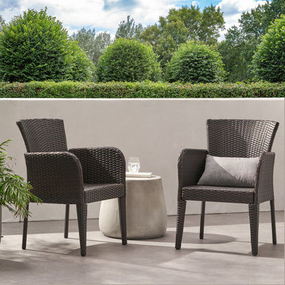 Two Anaya Outdoor Wicker Dining Armchairs with armrests and gray cushions are on a patio by a round table with a glass of water. Green bushes, trees, and a white wall stand in the background under partly cloudy skies.