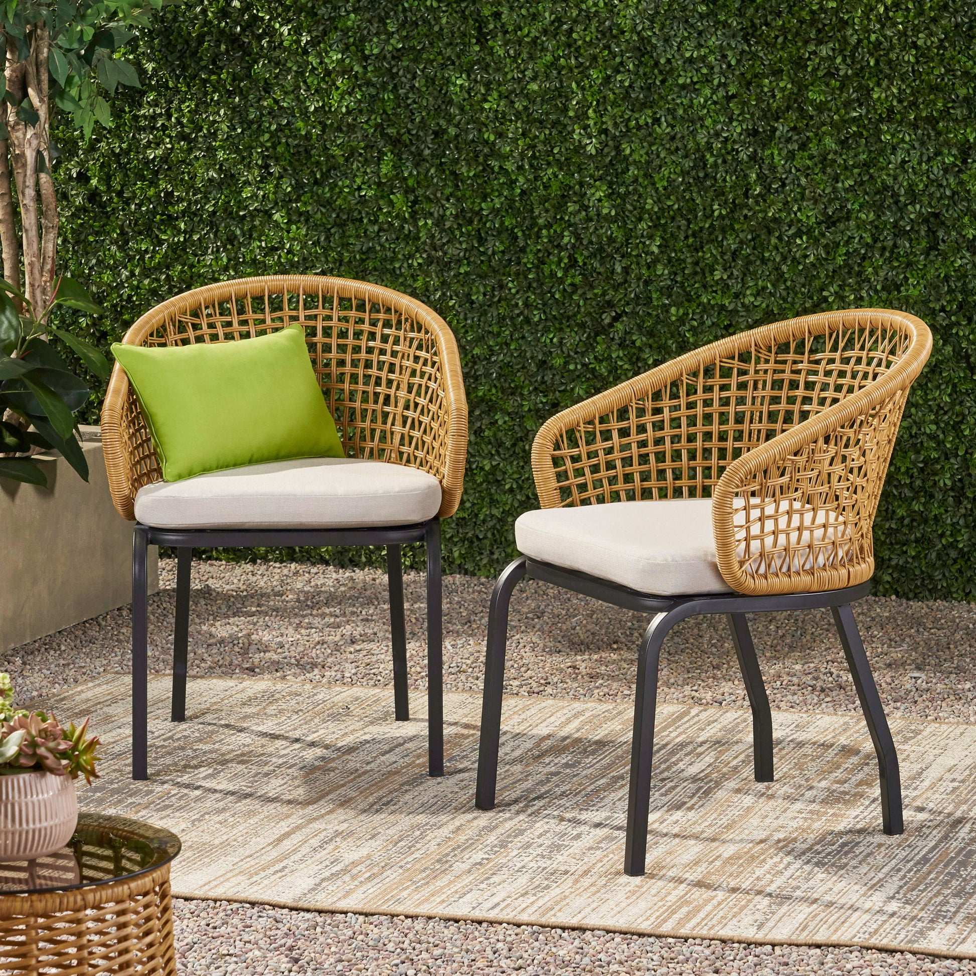 Two Outdoor Wicker Chairs With Powder Coated Iron Legs and light cushions sit on a striped outdoor rug over gravel, next to a potted plant. A green pillow accents one chair, with a leafy hedge in the background.