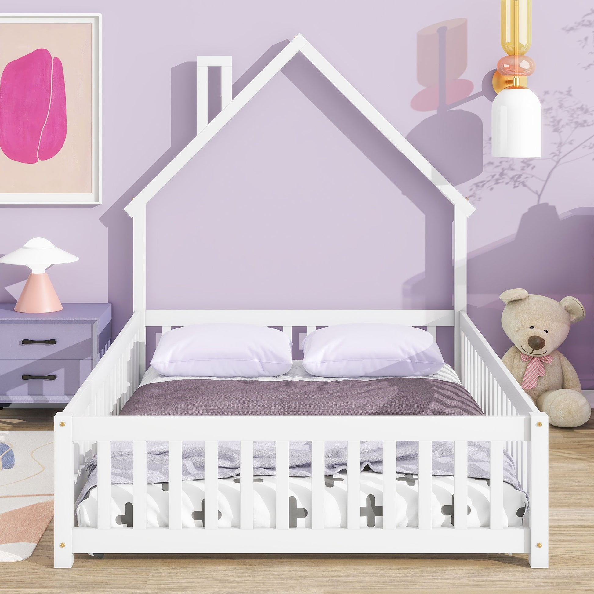 The House-Shaped Headboard Floor Bed With Fence in white stands against a light purple wall, dressed with pillows and a purple blanket. A teddy bear sits on the floor, while a nightstand with a lamp and abstract art are close by.