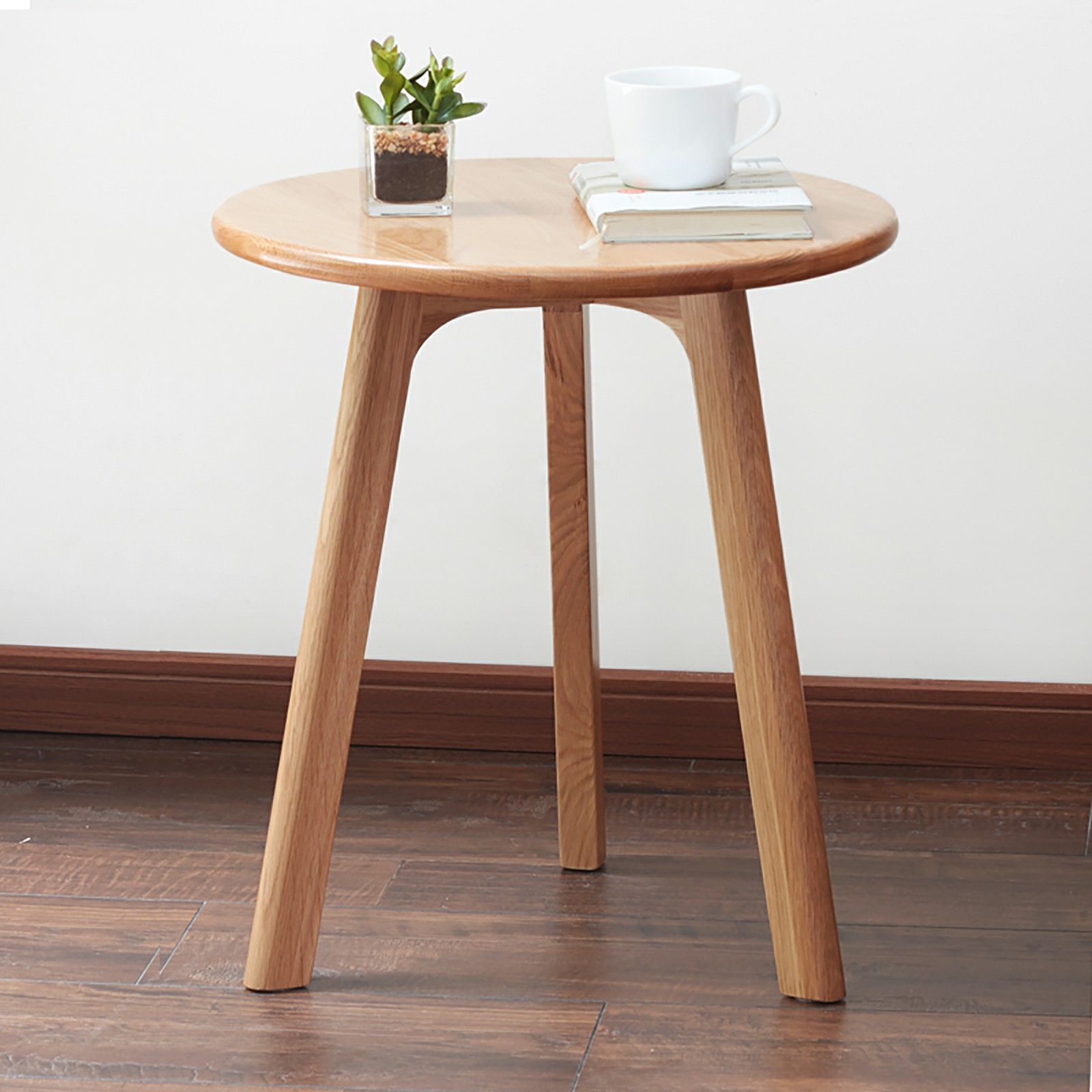 The Round Small End Table, perfect for bedroom or balcony, features three legs and a wooden top, holding a white cup, two stacked books, and a potted plant. It stands on a wooden floor against a plain white wall.