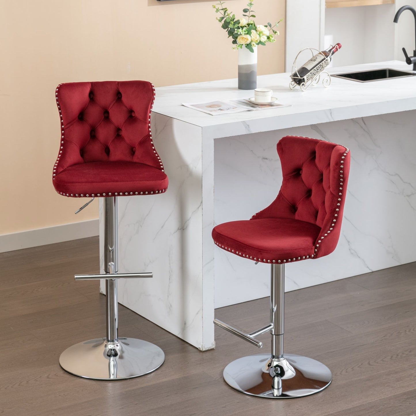 A set of two Swivel Velvet Barstools with tufted backs and chrome bases sits by a white marble kitchen island adorned with flowers, wine, and a glass, all atop a wooden floor. The stools feature adjustable seat heights from 25-33".