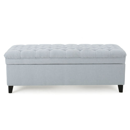 The Juliana Storage Ottoman features light gray upholstery, a tufted cushioned top, and dark birch legs, shown from the front.