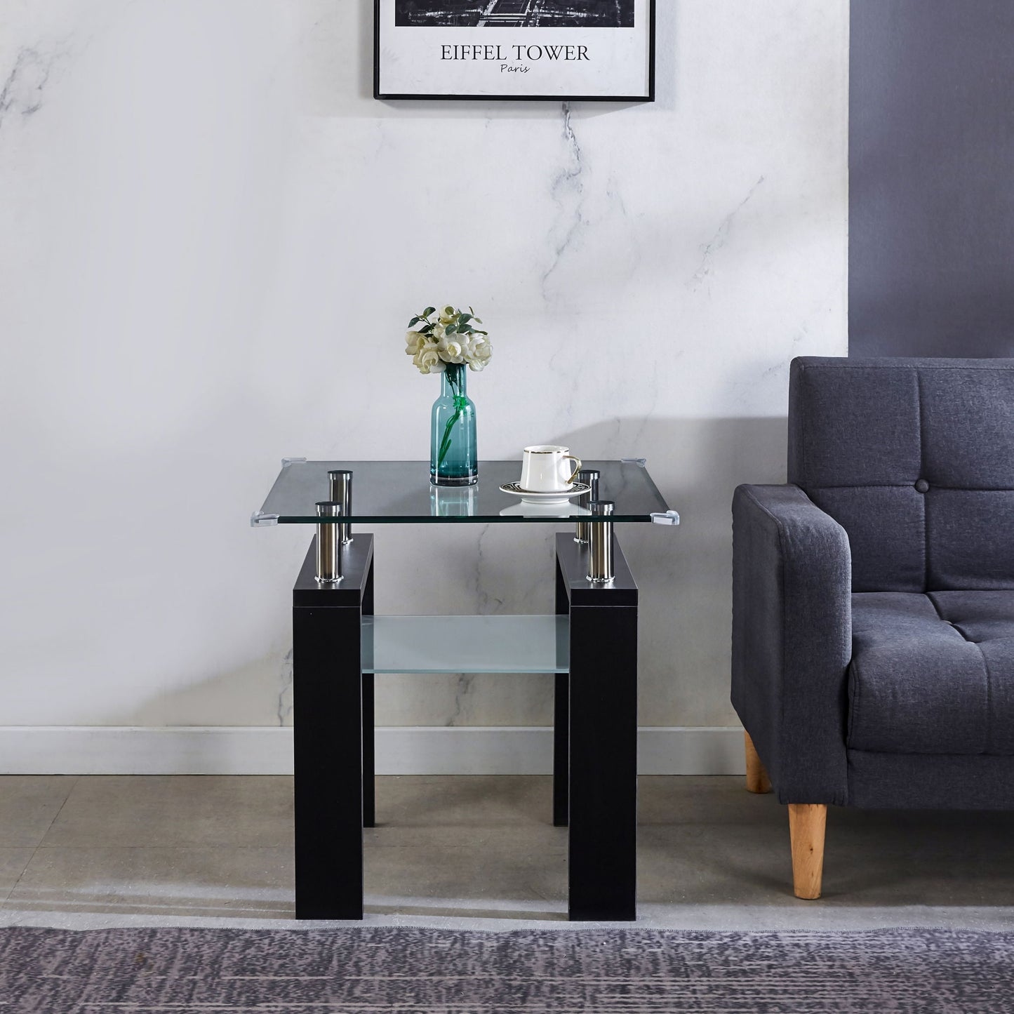 A modern living room features the Clear Glass Top Side Table with a green vase of white flowers and a white cup and saucer, placed beside a gray sofa and a framed picture on a marble-textured wall.
