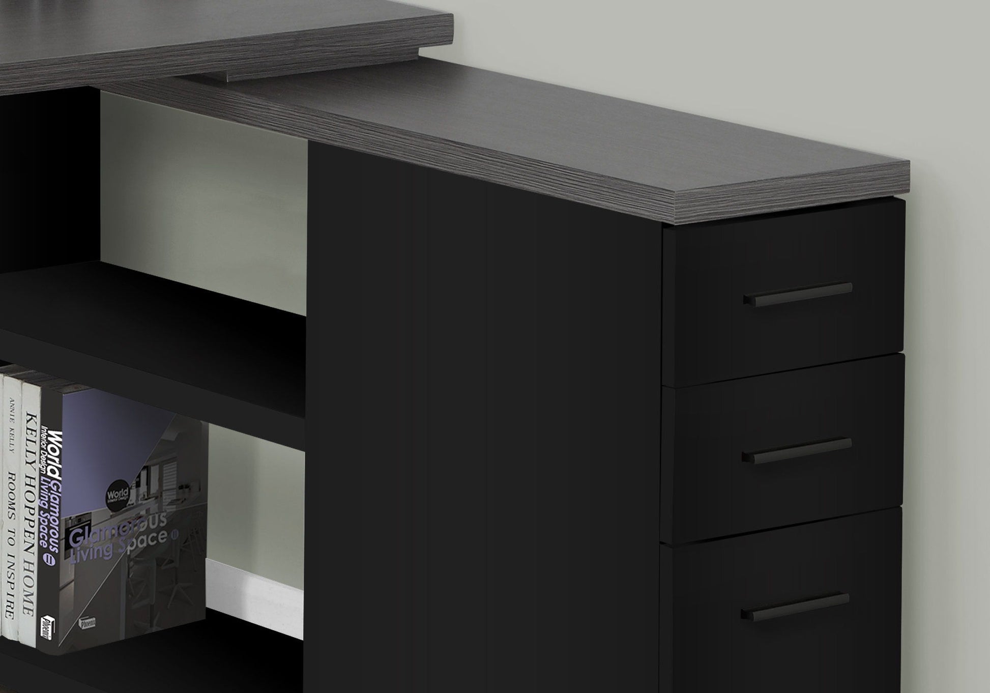 Close-up of a contemporary black L-shaped Computer Desk with storage drawers and a raised shelf, designed for home office use, featuring an open lower shelf with books, set against a light wall.