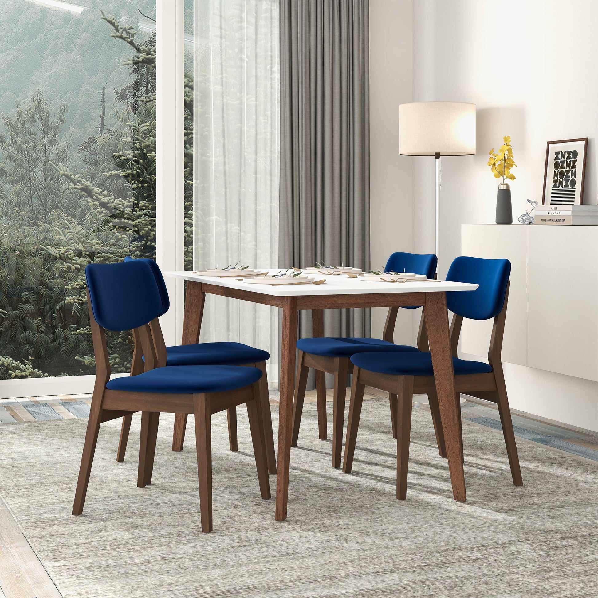 A modern dining room features a rectangular table, four Carlos - Solid Back Side Chairs with blue cushions, a floor lamp, a sideboard with decor, large windows with gray curtains, and tree views outside.