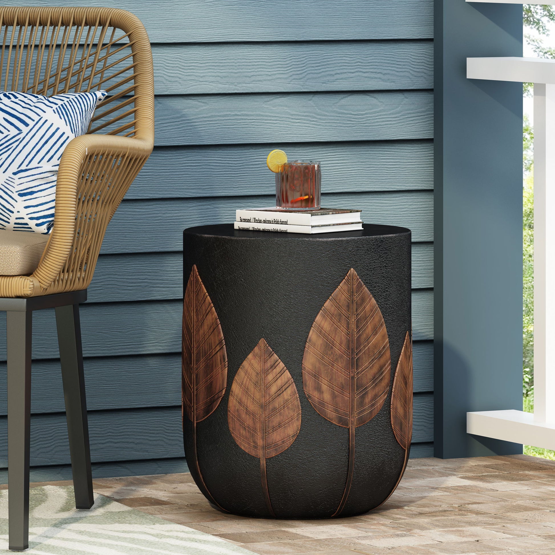The Outdoor Lightweight Concrete Side Table - Copper holds a drink and books on a porch beside a wicker chair with a blue-and-white pillow, set against blue siding and a white railing.