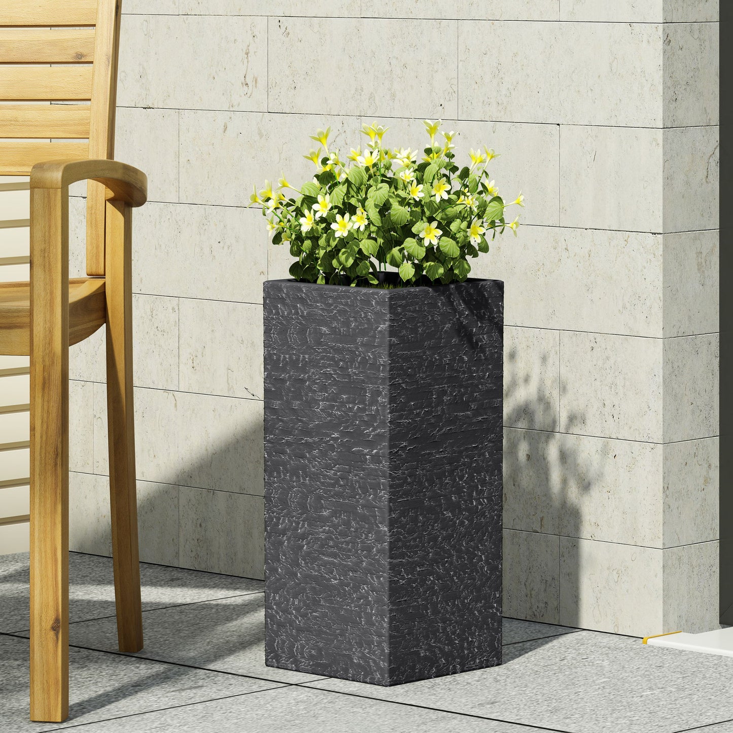 The Modern Outdoor Planter, a tall square black pot with yellow and white flowers, sits on a tiled patio beside a light wood chair and a wall of light beige stone tiles.