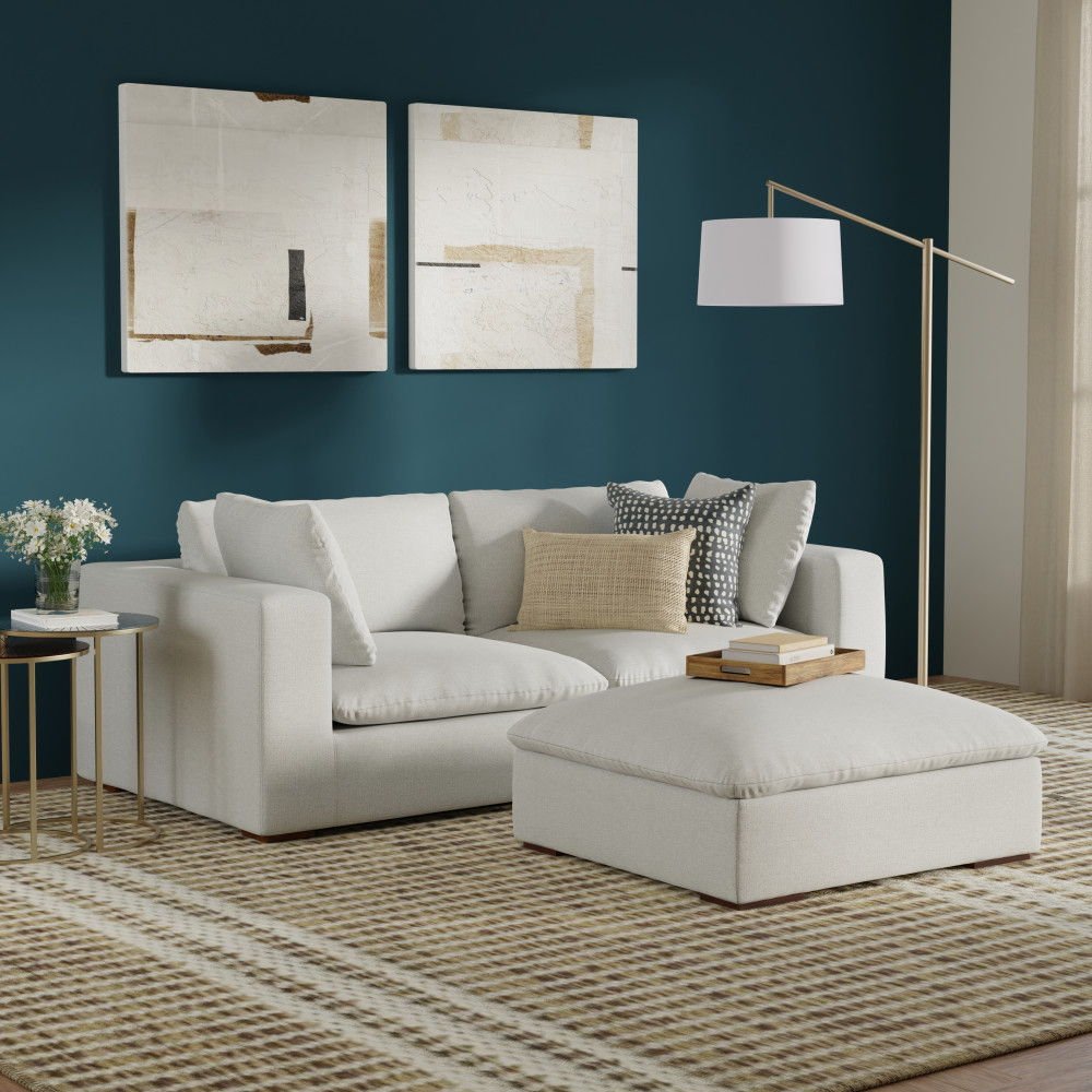 A modern living room with the Jasmine Handcrafted Sofa and Ottoman in cream, a patterned rug, side tables with flowers and books, a floor lamp, and two abstract art pieces on a dark teal wall. Natural light enters from the right.