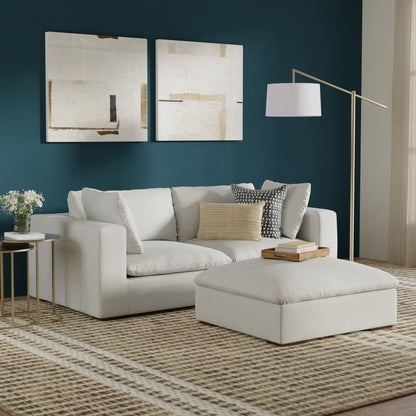 A modern living room with the Jasmine Handcrafted Sofa and Ottoman in cream, a patterned rug, side tables with flowers and books, a floor lamp, and two abstract art pieces on a dark teal wall. Natural light enters from the right.