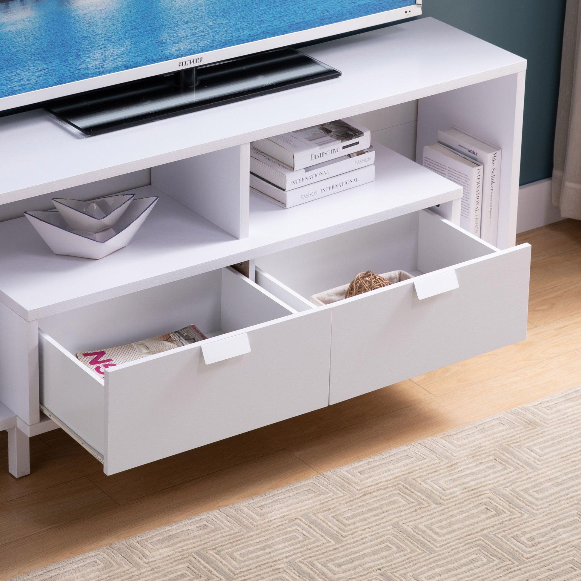 The Modern TV Stand & Two Shelves And Two Drawers features white finish, two open drawers for magazines and decor, shelves for books and bowls, all set on a wooden floor next to a beige rug.