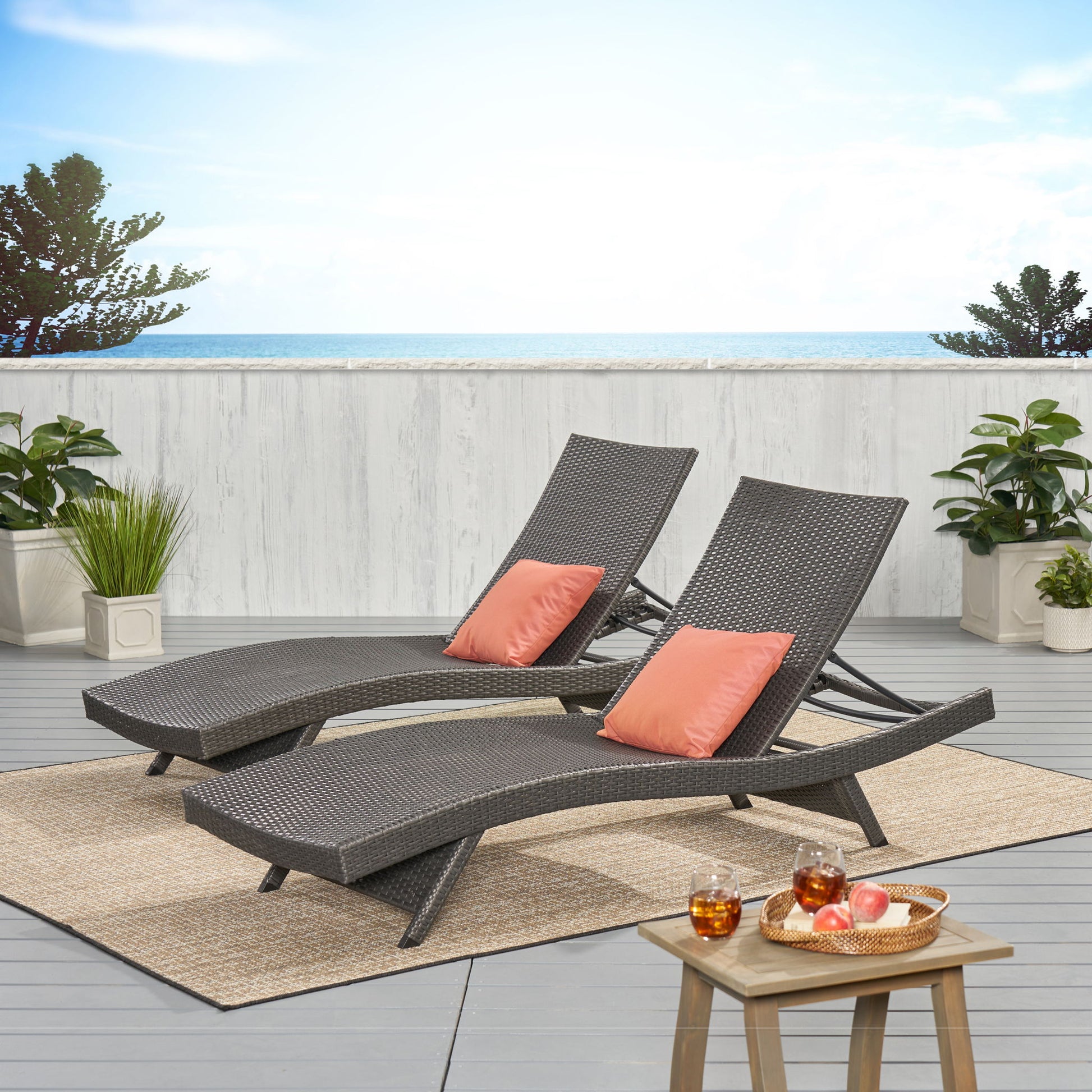 Two Salem - Chaise Lounge chairs with coral pillows rest on a patio rug facing the ocean. A small table nearby holds iced drinks and fruit, while potted plants and a white fence complete the patio setting.