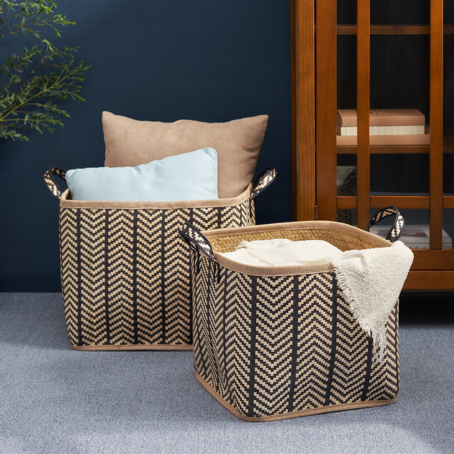 Two Square Palm Leaf Woven Wicker Storage Baskets with Handles (set of 2, black/brown) sit on a carpet, one holding two pillows and the other with a cream blanket. A wooden cabinet and green plant are visible in the background.