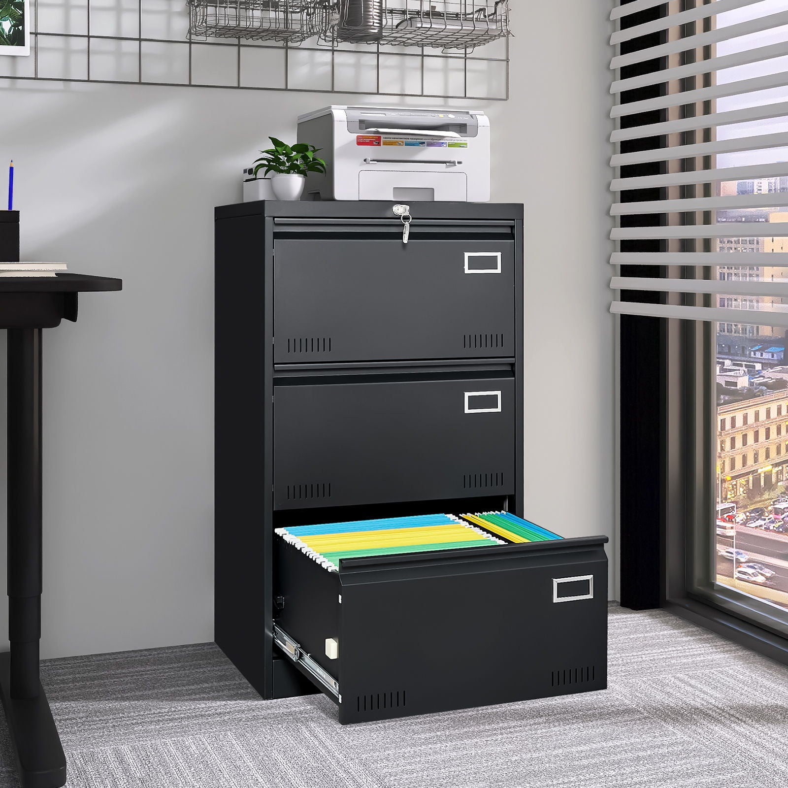 A Filing Cabinet Lateral File Cabinet 3 Drawer sits by a window with the bottom drawer open, revealing colorful files. A printer and plant rest on top, while sunlight filters through horizontal blinds onto office carpeting.