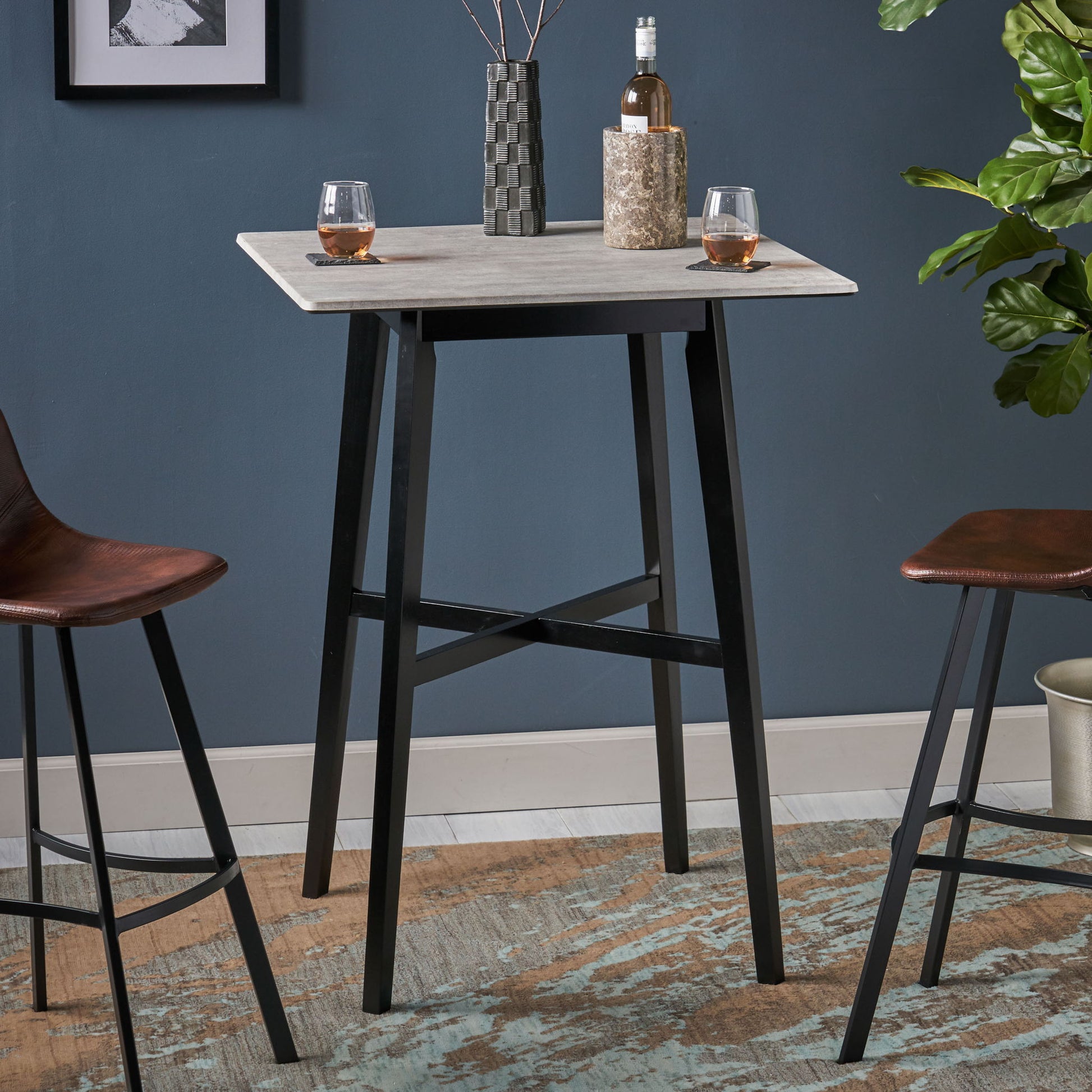 The Modern Bar Height Dining Table with a light laminate top and rubberwood legs stands between two brown barstools, holding a vase, a bottle, and two glasses. A plant and framed artwork are visible in the background.