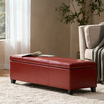 The Glouster - Contemporary Storage Ottoman in red leather sits on a light rug beside a beige sofa with a green throw, while sunlight streams through the window and a potted plant decorates the background.