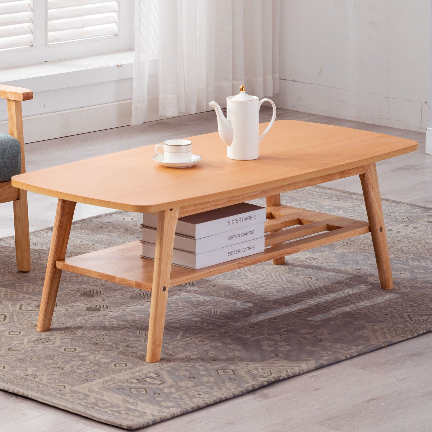 The Bahamas - Coffee Table features angled legs, a lower shelf with three stacked books, and a top surface holding a white teapot and cup. It rests on a patterned rug in a sunlit room with sheer curtains and a window.
