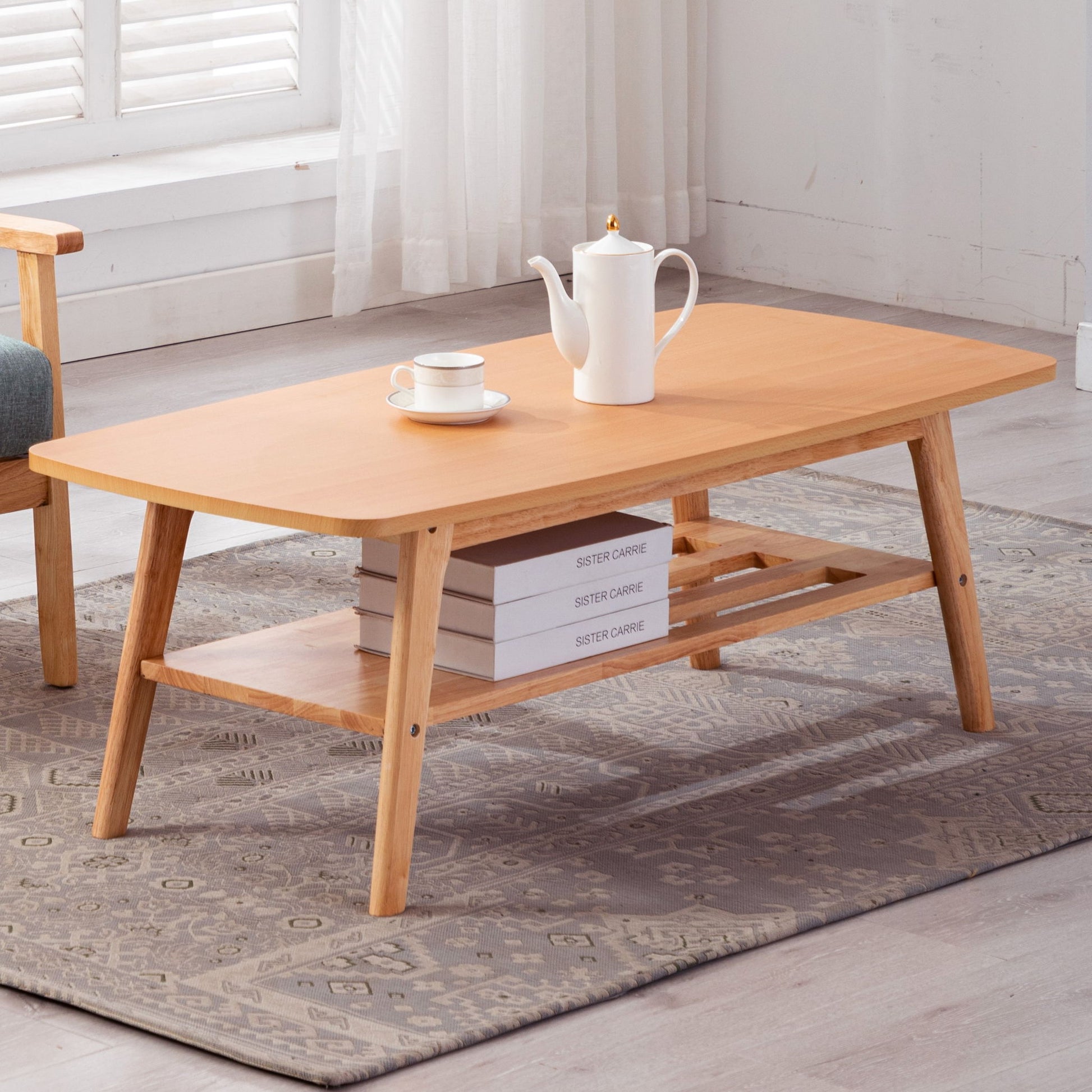 The Bahamas - Coffee Table features angled legs, a lower shelf with three stacked books, and a top surface holding a white teapot and cup. It rests on a patterned rug in a sunlit room with sheer curtains and a window.