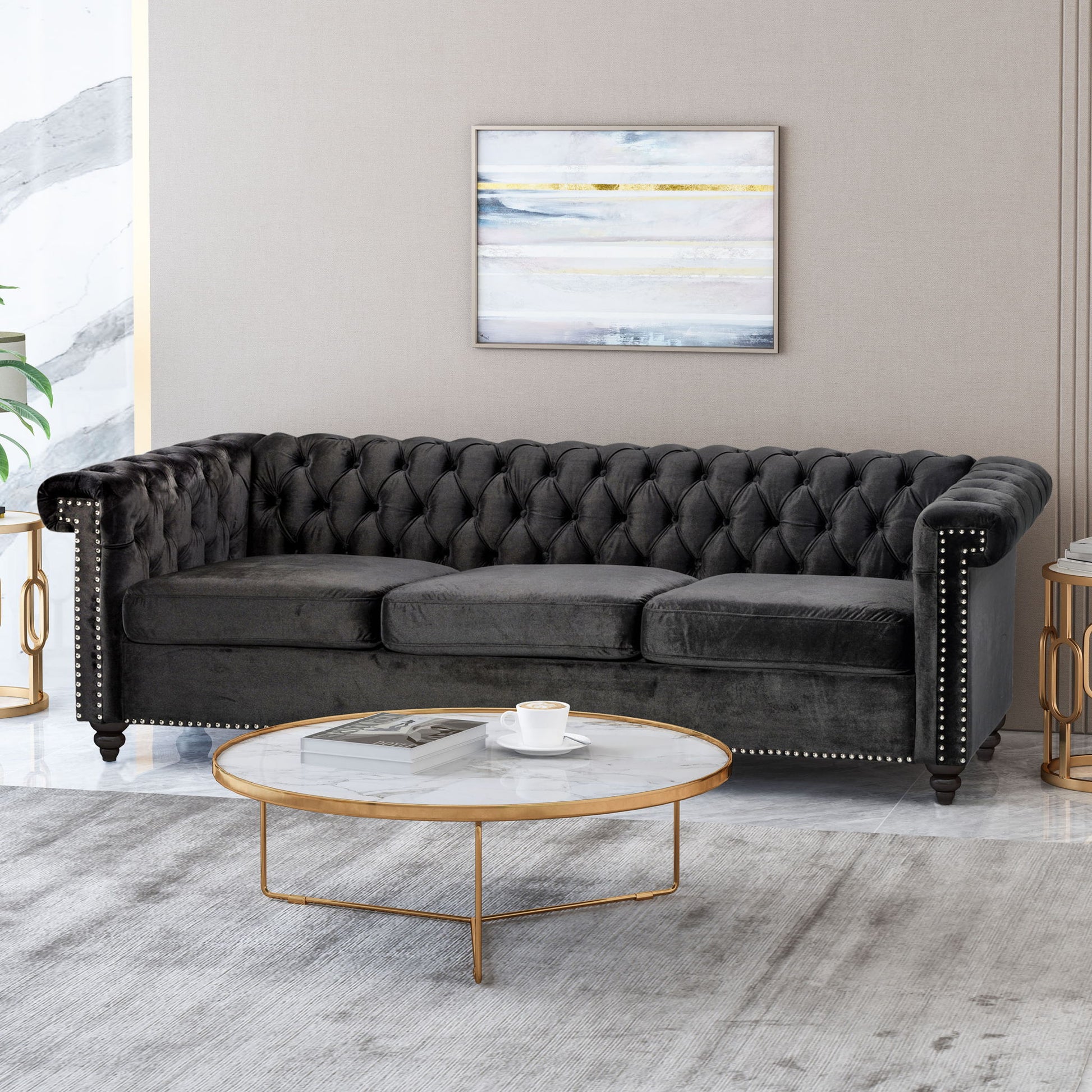 A modern living room features a Chesterfield Style 3 Seater Sofa with deep button tufting, a round marble and gold coffee table with a cup and saucer, a light gray rug, and a framed abstract painting on the wall.