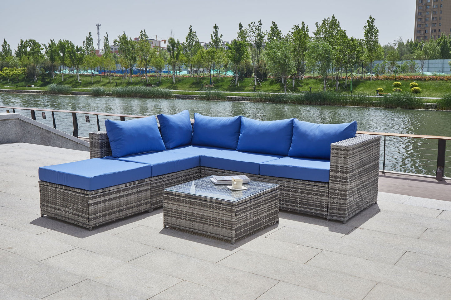 A Patio Sofa With Cushions featuring gray wicker and blue cushions is set around a glass-top coffee table on a stone terrace that overlooks a tree-lined river.
